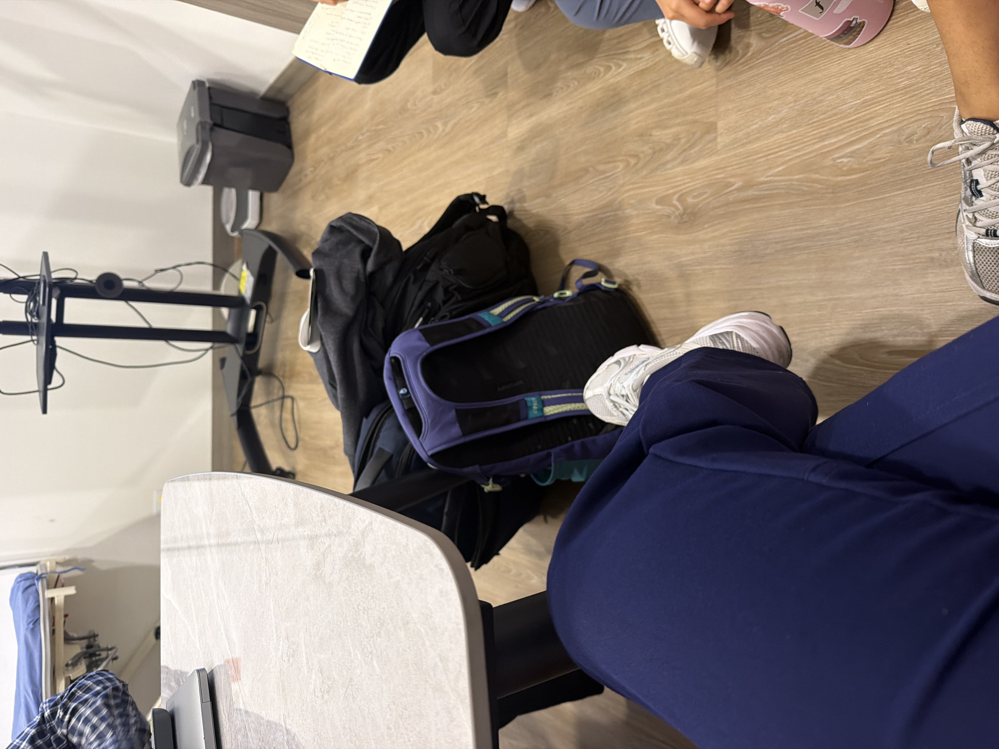 The image, taken from a slightly elevated, rotated perspective looking down, shows the lower bodies of at least five individuals gathered indoors. The setting features a light-colored wooden floor.
In the foreground, a person's legs, clad in navy blue scrub-like pants, are visible, along with white and grey athletic shoes. Next to them, a black backpack and a distinctive purple and blue backpack (possibly with "LIMA" text and a logo on a strap) are placed on the floor, alongside what appears to be a grey hooded garment.
Around the bags, other people's feet and lower legs are visible. To the right, another pair of white and grey athletic shoes, and further up, a person wearing light blue/grey pants with white athletic shoes. Adjacent to these are pink slip-on shoes adorned with several small stickers. In the upper left corner, a person wearing black pants holds an open notebook with handwritten text, indicating some form of discussion or study.
Further objects in the scene include a dark grey electronic device mounted on a wall or stand in the background, and a black metal stand with cables, suggesting the presence of technical equipment. A light-colored desk or table edge is visible in the bottom-left, and in the far background, a clothes rack with hanging blue fabric and a plaid shirt can be seen.
The people appear to be sitting on the floor or very low furniture, engaged in an activity that involves note-taking. The visible text on the notebook is handwritten notes, mostly illegible, but some English words can be vaguely perceived. The overall atmosphere suggests an informal meeting, study session, or break time in an indoor environment, possibly an office, clinic, or educational setting. The location is specified as Bojacá, Colombia, though no specific visual cues within the image confirm this.