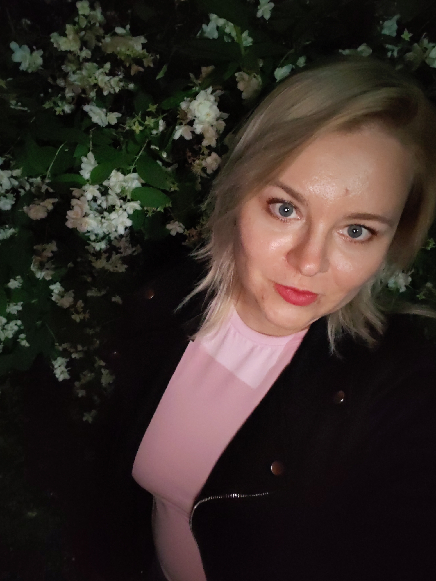 The image shows a close-up selfie of a blonde woman with blue eyes and red lipstick. She is wearing a pink shirt and a black jacket. She is standing in front of a bush with white flowers. The image is dark and appears to have been taken at night. There is no visible information in the image that would allow for the identification of a specific location within the city of Izhevsk, Russia.