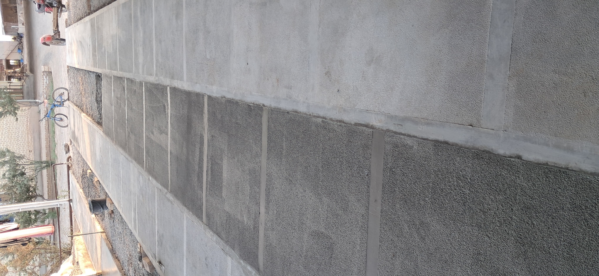 The image shows a ground-level view of a paved area, possibly a walkway or parking lot, in Mary, Turkmenistan. The paving is composed of large, rectangular concrete slabs with visible seams between them. Some of the slabs have a rough, pebbled texture, while others appear smoother. A narrow strip of gravel runs alongside the paved area.
In the background, on the left side of the image, a blue bicycle is leaning against something. Further back, a motorcycle is parked. There are also glimpses of buildings and trees, suggesting an outdoor setting with some signs of development or construction. The lighting indicates it is either early morning or late afternoon. The weather appears to be clear.
The scene appears to be a quiet, perhaps residential or communal area. There are no people clearly visible in the frame. The overall impression is one of a functional, outdoor space. No text is visible in the image.