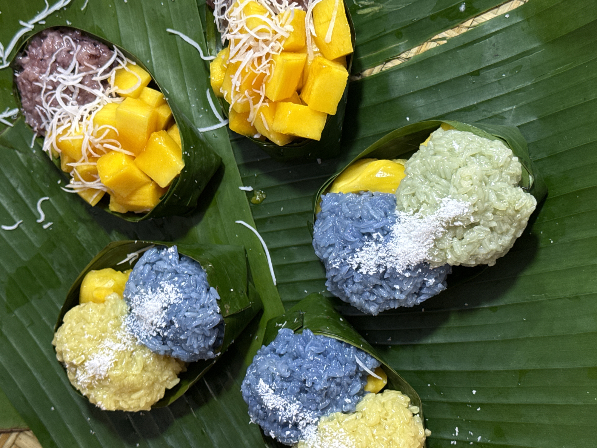 Four small banana leaf parcels filled with colorful sticky rice and diced mango are arranged on a bed of fresh green banana leaves. The parcels contain combinations of yellow, blue, and light green sticky rice, with some parcels also featuring purple sticky rice. All are topped with shredded coconut. The scene appears to be an outdoor setting in Nanshan District, China, suggested by the presence of fresh produce and natural elements, possibly at a local market or a street food vendor.