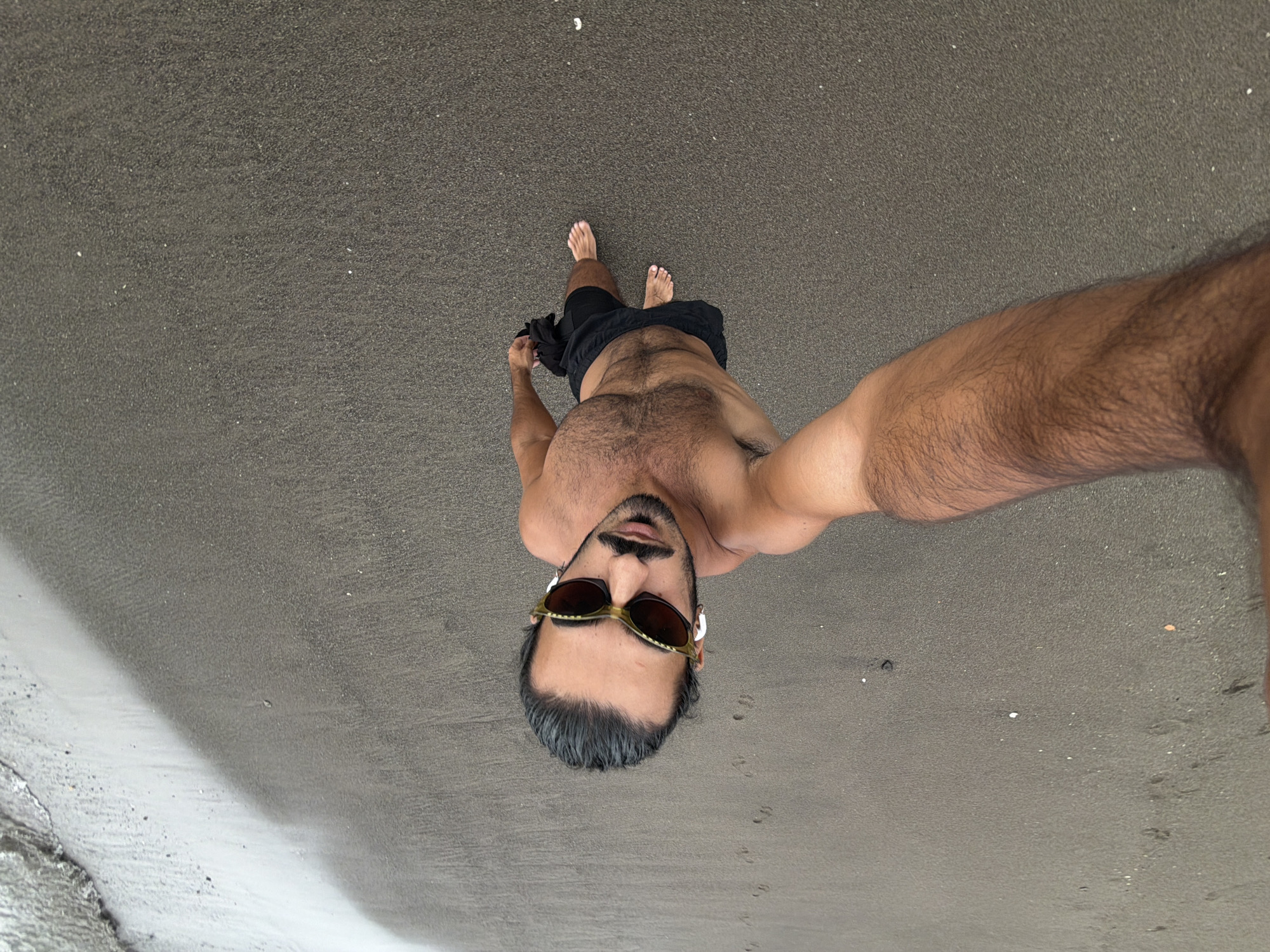 A person is standing on a wet, sandy beach, with the water edge visible to the upper left. The person is wearing black swim trunks and is shirtless, with sunglasses on. Their right arm is extended towards the camera, holding what appears to be a black cloth. The sandy surface shows a few footprints. The lighting suggests it is daytime. The specific location in Mengwi, Indonesia is not identifiable from the image.