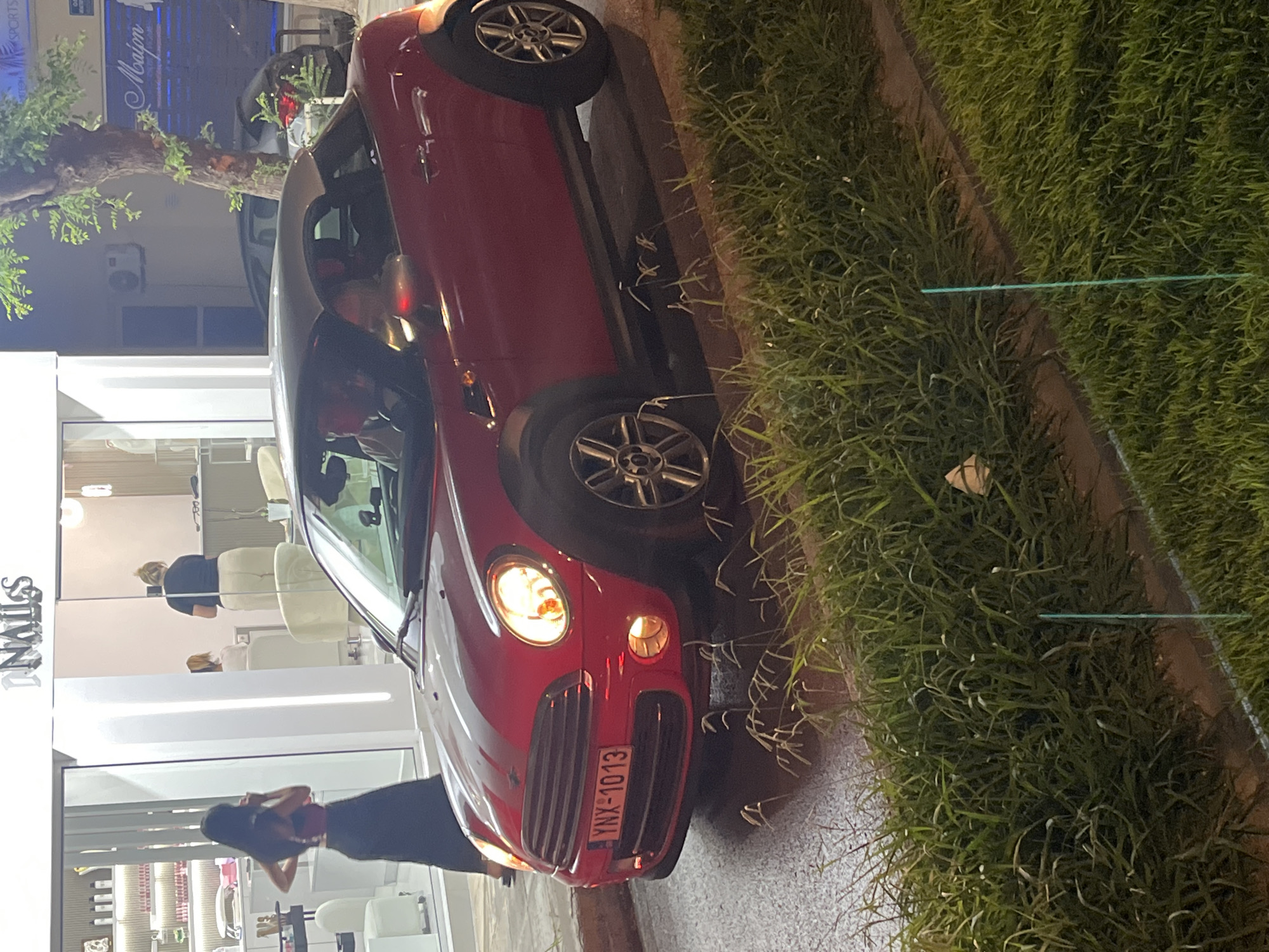 A red Mini Cooper coupe with its headlights illuminated is parked on a street in Athens, Greece, in front of a nail salon. The license plate reads "YNX-1013". A woman with dark hair is standing on the sidewalk to the left of the car, facing away from the camera and talking on her phone. Inside the salon, two women are visible; one appears to be a client sitting in a chair, and another woman, likely an employee, is standing. The salon has a modern aesthetic with white walls and displays of nail products. To the right of the salon, another business with a sign reading "Major" is visible. The street is lined with trees and greenery. The scene is captured at night.