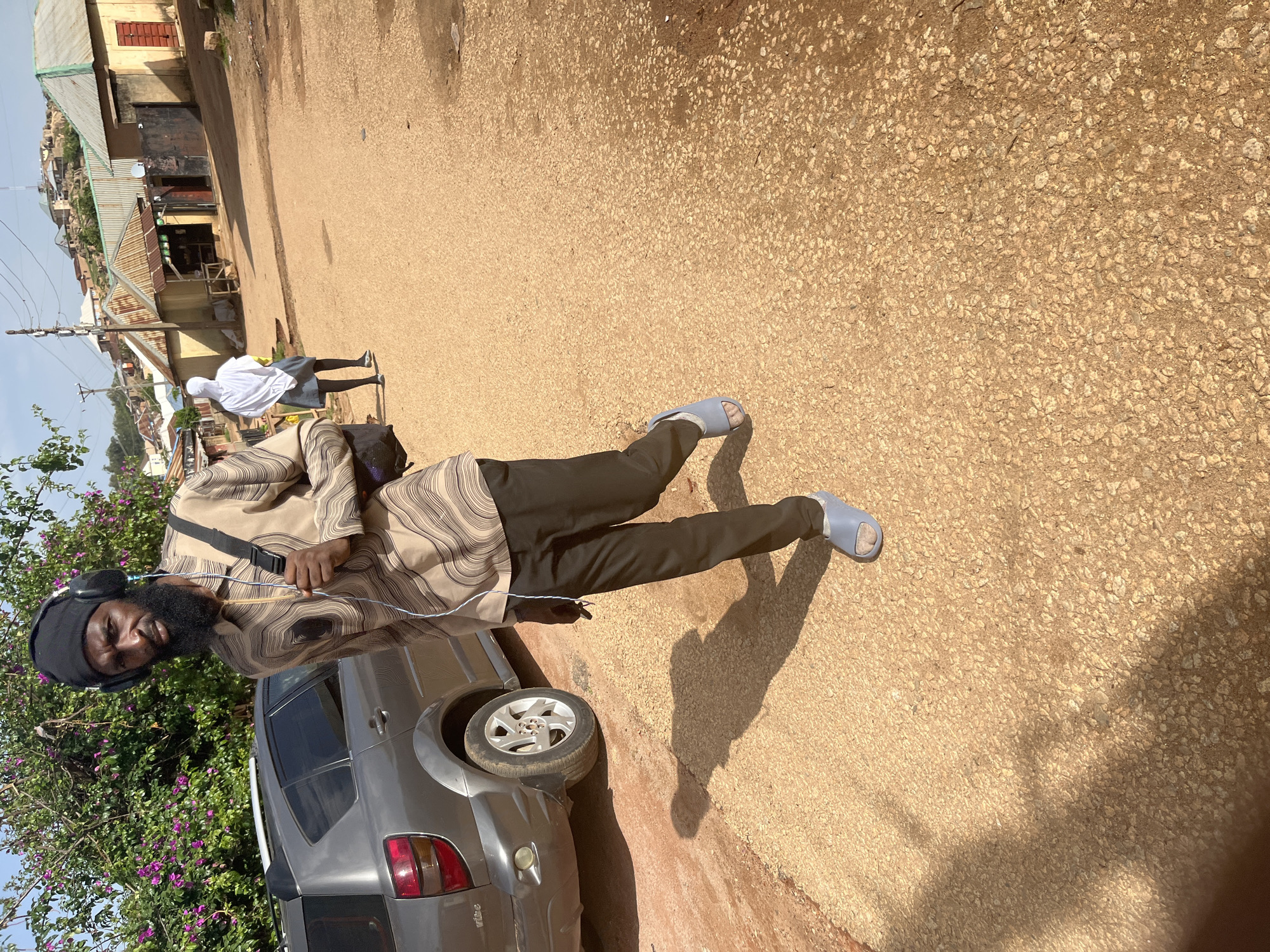 The media file shows a man standing outdoors on a dirt road. He is wearing a patterned robe, dark pants, a black head covering, and headphones. He has a beard and appears to be holding a wooden stick. To his left, a grey car is parked. In the background, there are buildings with corrugated metal roofs and a hill. To the left of the man and the car, there are flowering bushes. The scene appears to be a residential or commercial area in Jos, Nigeria. The sun is shining, casting a shadow from the man on the ground, suggesting it is daytime. Another person, partially visible and wearing a white headscarf and blue skirt, is walking in the background. The overall impression is a candid shot of daily life in a Nigerian town.
