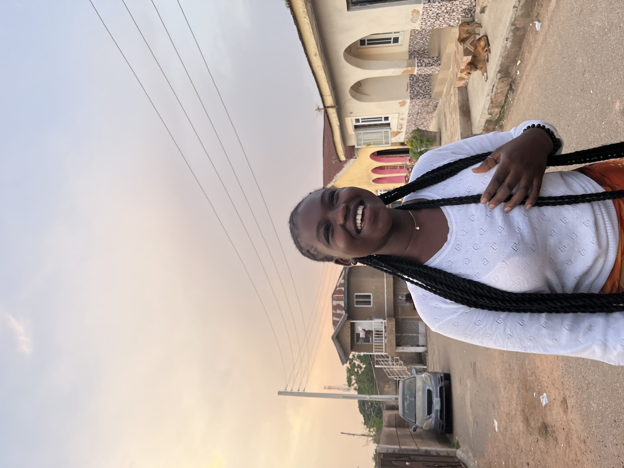 A young woman with dark skin and long black braided hair is standing on a dirt road, smiling at the camera. She is wearing a white long-sleeved shirt with a subtle patterned texture and an orange skirt. Her right hand is placed on her chest. The background shows a residential area with buildings and a street. To the left of the woman, a grey sedan is parked. In the middle ground, a two-story building with a balcony and stairs is visible. To the right, another building with arched doorways is seen, and two dogs are resting on the concrete steps. The sky is a gradient of soft orange and blue, suggesting either sunrise or sunset. The scene appears to be in a neighborhood in the city of Gura Top, Nigeria.