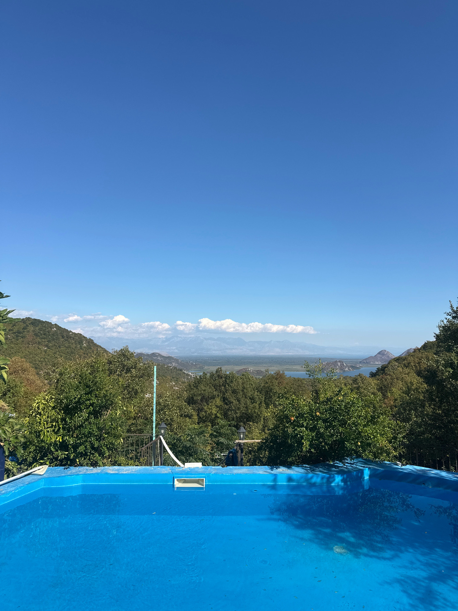 A bright blue swimming pool occupies the foreground of this image, reflecting the surrounding greenery and sky. Beyond the pool, a verdant landscape unfolds, characterized by rolling hills covered in dense trees. In the distance, a body of water, likely a lake, is visible, with mountains forming a backdrop under a clear blue sky. The scene is set outdoors in a location that appears to be in or around Cetinje, Montenegro, a region known for its natural beauty. The photograph captures a serene and scenic view, likely taken from a vantage point overlooking the landscape.