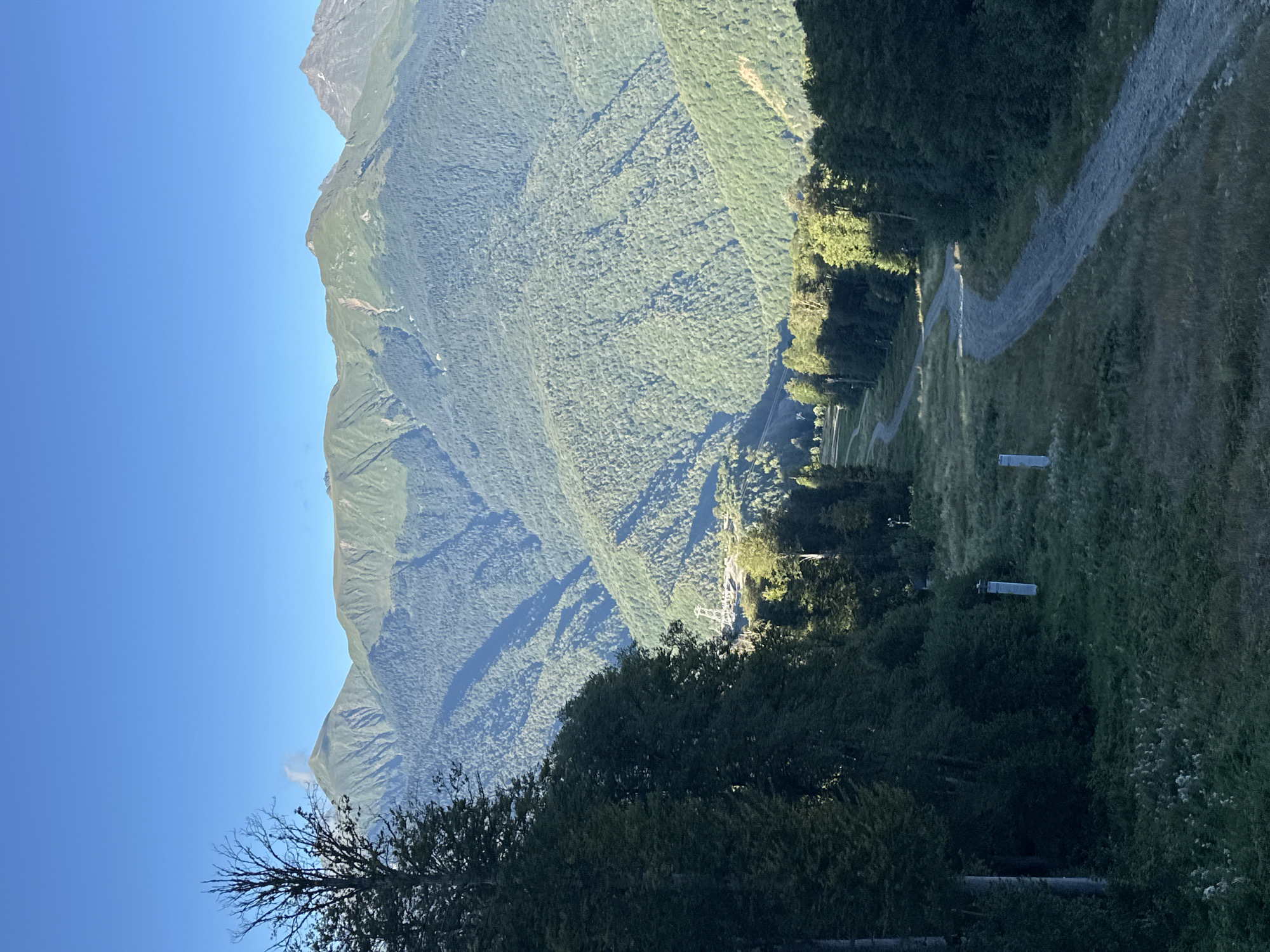 The image depicts a mountain landscape in Estosadok, Russia, with a clear blue sky. In the foreground, a winding dirt path curves through grassy slopes bordered by dark green trees on the left and right. The path appears to be a ski trail or hiking path. In the mid-ground, more trees and the start of steeper mountain slopes covered in dense forest are visible. Further in the distance, layers of mountains rise, with the highest peaks showing rocky, lighter-colored summits and shadowed valleys. A ski lift structure is visible in the mid-ground, nestled amongst the trees. The overall scene is one of natural beauty and outdoor recreation.