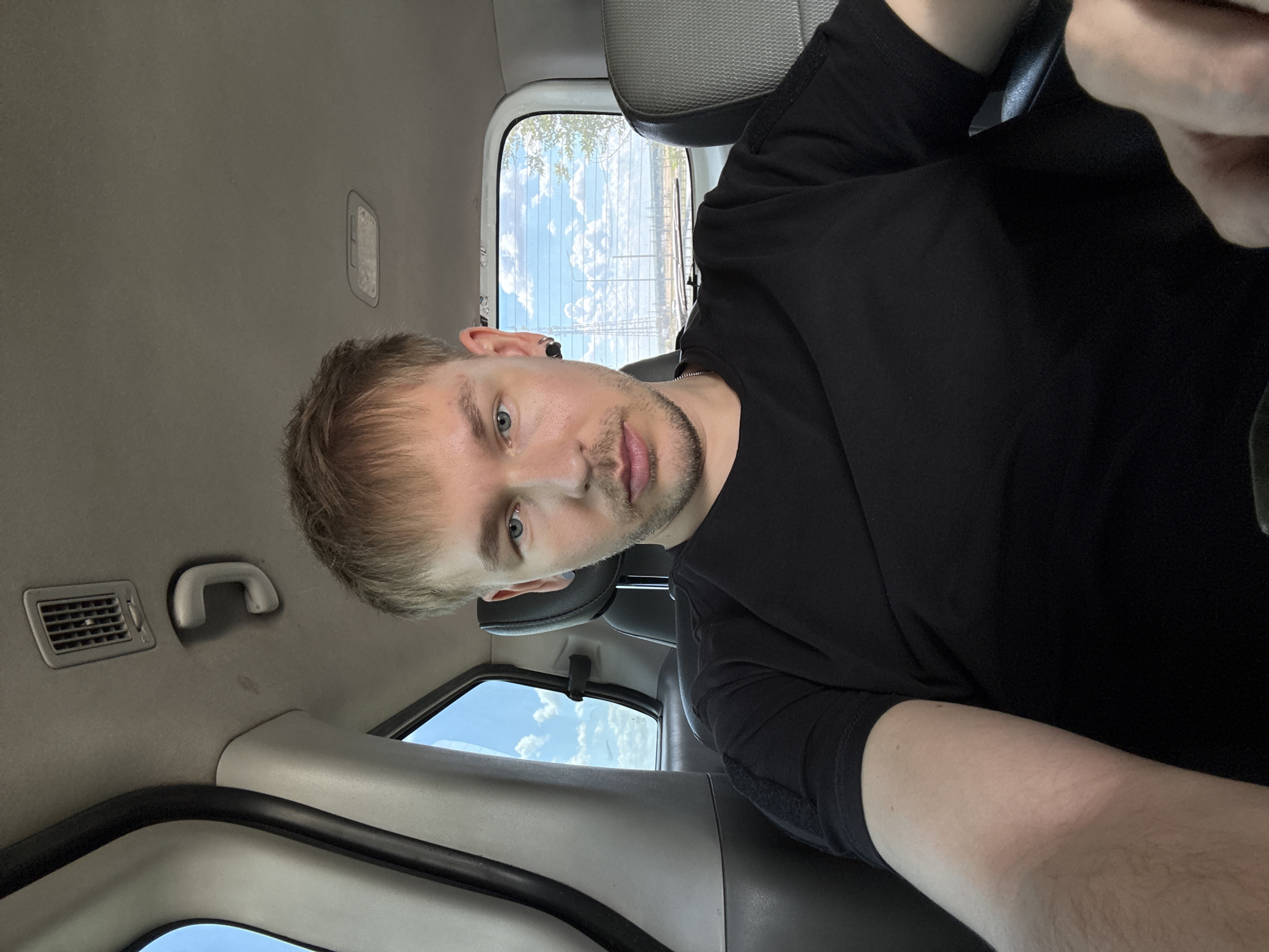 A young adult male with blonde hair and blue eyes is seated in the back of a vehicle, possibly an SUV. He is wearing a black t-shirt and has a black ear gauge in his left ear. The interior of the vehicle shows gray headliner with a vent and a grab handle. The window behind him reveals a bright, sunny sky with scattered clouds, and a landscape with trees and what appears to be power lines or a fence in the distance. The overall setting suggests a daytime scene in a rural or suburban area. The location is Pervomaiskii, Russia.
