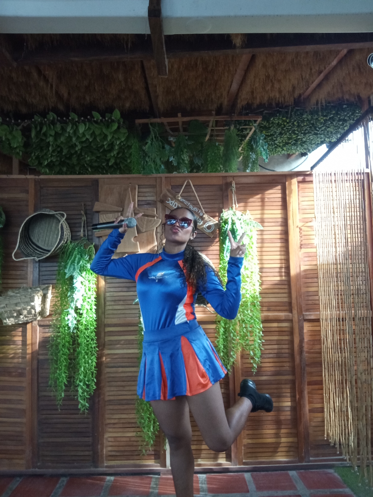 A woman is captured in a dynamic, full-body shot, striking a playful pose in what appears to be a covered, rustic-themed venue. She is wearing a long-sleeved top and a pleated mini-skirt, both in a striking combination of royal blue and orange. Dark, reflective sunglasses adorn her face, and her lips are pursed in a playful pout. Her dark, curly hair falls over her shoulder. She holds a silver microphone in her left hand, while her right hand is raised to display a peace sign. One leg is lifted, and she wears black sneakers.
The background consists of a wall made of light-brown wooden slats, adorned with various decorative items. These include several woven baskets of different sizes and shapes, and other wooden carvings or decorative pieces. Lush green artificial plants hang profusely from the ceiling and are draped along the walls, adding a vibrant touch. The ceiling above is a thatched roof supported by visible wooden beams, with a white structure bordering the top of the frame. To the far right, a vertical screen made of thin, light-colored bamboo or wooden reeds provides additional texture. The floor beneath her is made of reddish-brown tiles, laid in a pattern with visible grout lines.
A small, light-colored wooden sign is hanging on the wall behind the woman's head; the text on it is mirrored and partially legible, appearing to spell "comprasuqui..." or similar. An indistinct logo is also visible on the right shoulder of her top. The setting, with its natural materials and abundant greenery, suggests a tropical or resort-like ambiance, consistent with Porlamar, Venezuela. The woman's pose and expression convey a sense of confidence and fun.