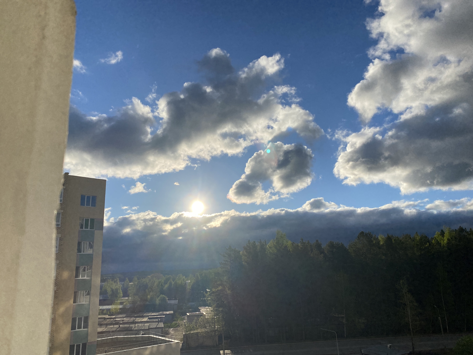 This image shows a view from a window in Chaikovsky, Russia. The sky is mostly blue with numerous white and gray clouds. The sun is visible behind the clouds, casting rays of light downwards. Below the clouds, there is a line of trees, darker in color, possibly a forest or a large park. Beyond the trees, there are several low-rise buildings and a glimpse of the city landscape. On the left side of the frame, the edge of a tall building is visible, including windows. The photo is taken during daylight.