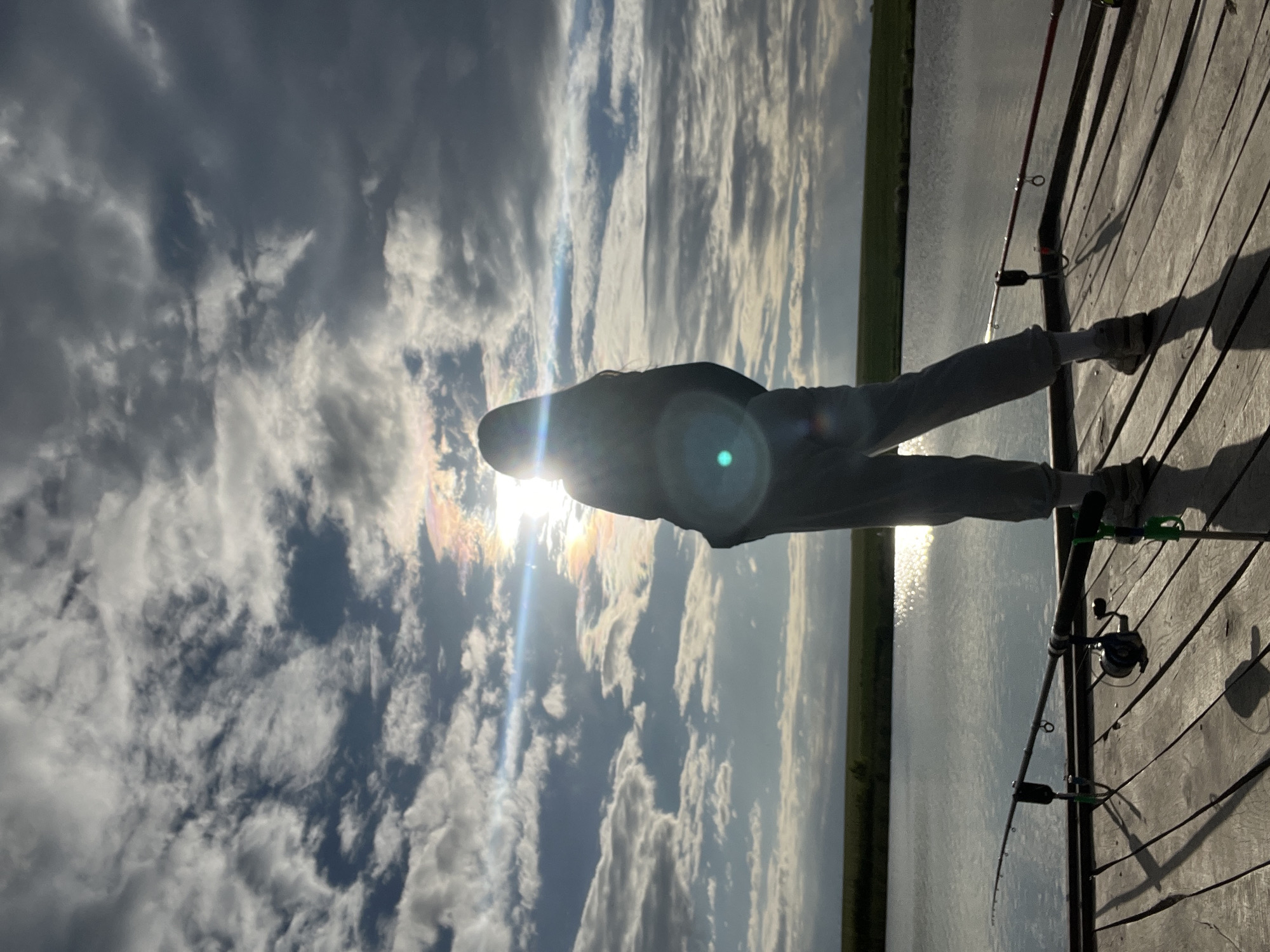 A person stands on a wooden pier against the setting sun. Fishing rods rest against the pier. Water reflects the sunlight. Clouds are in the sky. The location of this image could be Rudky, Ukraine.