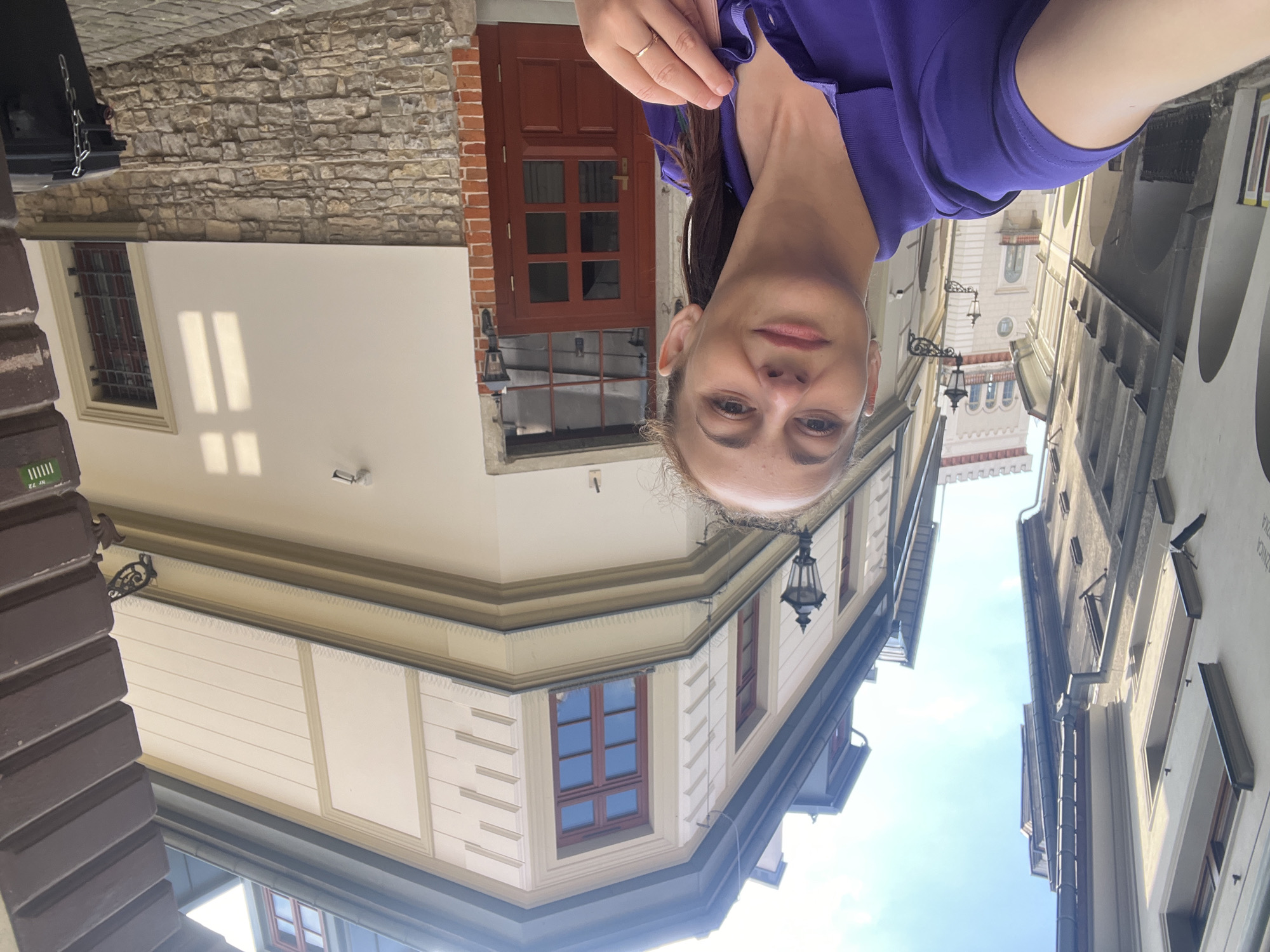 A woman is taking a selfie in front of a building on a cobblestone street. She is wearing a purple polo shirt and has her hair in a ponytail. The building behind her is a light-colored, two-story structure with a red door and a stone base. There is a lamppost on either side of the entrance. In the background, there are other buildings lining the street, creating a narrow urban canyon. The sky is blue with some clouds.