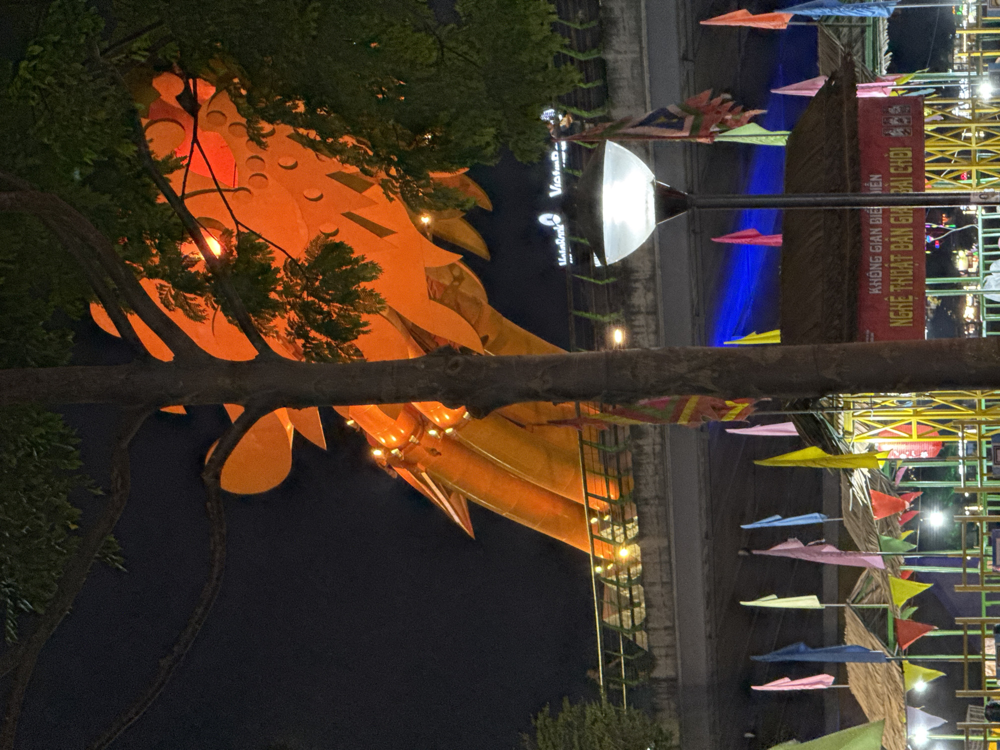 At night, the image showcases a partial view of the Dragon Bridge in Đà Nẵng, Vietnam. The bridge's orange dragon head is visible through tree branches. A lamppost with a white light stands to the right of the dragon. Across the river, a building with the words "VietinBank" displayed vertically can be seen. Festival decorations, including colorful pennants and a structure, are also present.