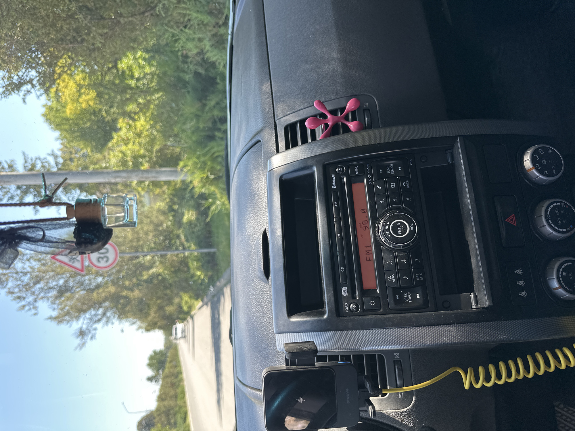 The image shows the interior of a car dashboard, looking forward through the windshield on a sunny day. The dashboard is dark gray plastic with a central console. A car stereo is visible, displaying "FM1 90.0". To the left of the stereo, a black smartphone mount is attached to an air vent, holding a black smartphone. A bright yellow coiled cable extends from the phone mount. A pink, stick-figure-like air freshener is attached to the right side of another air vent.
Through the windshield, a paved road is visible, curving to the left. A white car is driving in the distance on this road. On the right side of the road, there are trees and bushes. A road sign showing "30" within a red circle is visible on the left side of the road. The sky is clear and blue. The overall scene suggests a view from the driver's or passenger's seat of a car in Makiv, Ukraine, as indicated by the potential location.
