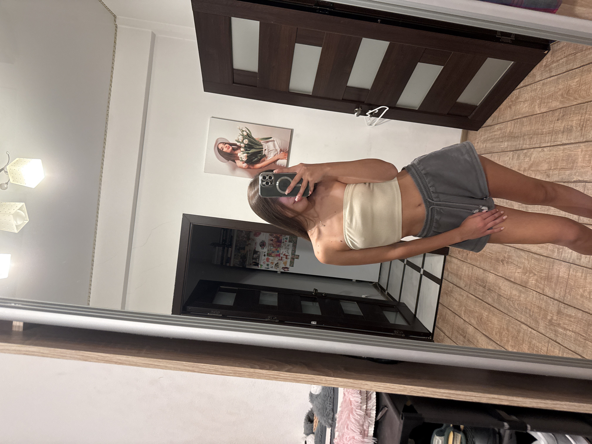 A young woman is taking a mirror selfie in what appears to be a bedroom or dressing area. She is wearing a light-colored, strapless crop top and grey athletic shorts. She is holding a smartphone in her right hand to capture the image. The background includes a large mirror, a wooden-framed door with glass panels, a painting of a woman holding flowers on the wall, and a tiled floor leading into a hallway. The room is brightly lit by ceiling lights. The style of the decor, particularly the doors and the artwork, suggests a contemporary residential interior. Without specific landmarks, it is impossible to determine the exact location within Chișinău, Moldova.