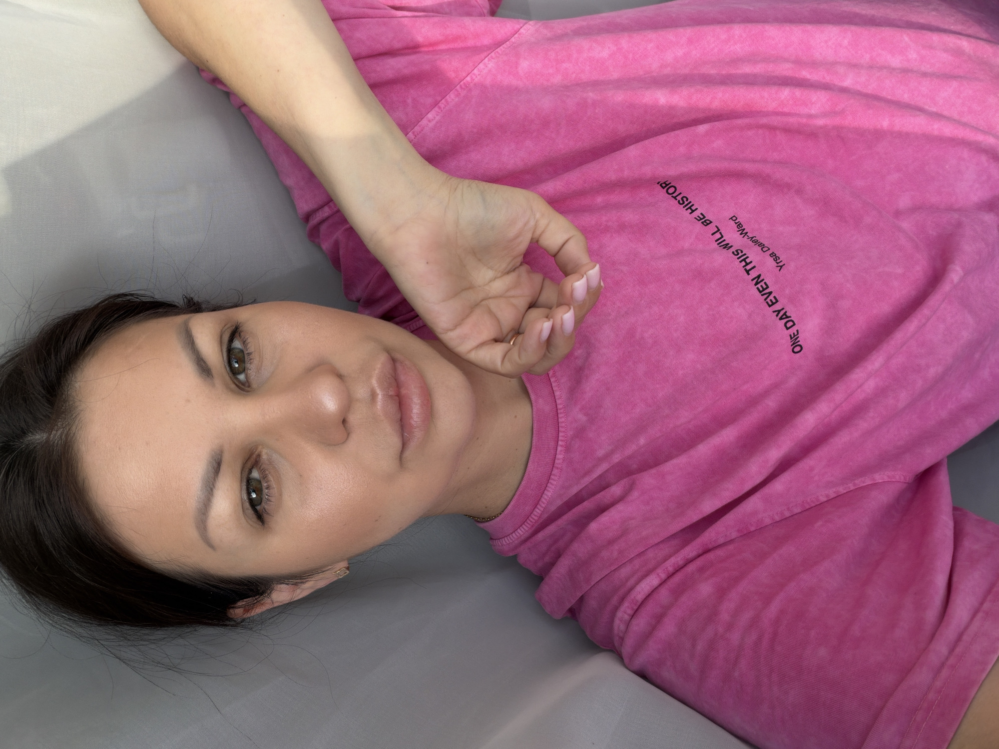 The image shows a close-up, overhead, outdoor selfie of a woman with brown hair and green eyes. She is wearing a pink t-shirt and a gold necklace. She is lying down and her right arm is bent, with her hand resting on her chest. The background is a light gray curtain. There is no indication that the image was taken in Sochi, Russia.