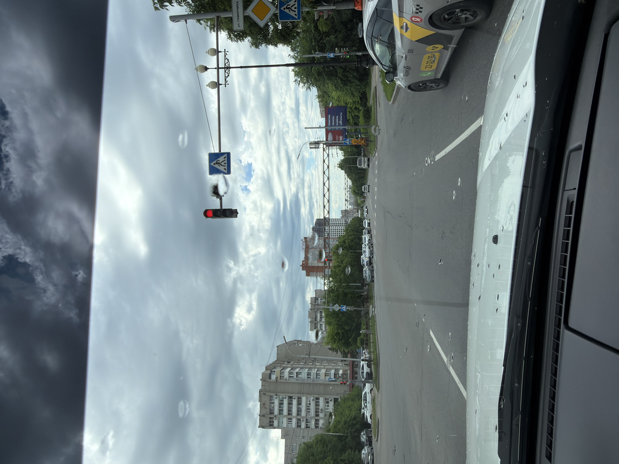 The image depicts a view from inside a vehicle looking out through the windshield at an urban intersection in Khabarovsk, Russia. The sky is overcast with dark clouds, and there are several water droplets visible on the windshield. The road ahead shows a traffic light that is red, indicating a stop. A pedestrian crossing sign is visible on the left side of the intersection. Several buildings, appearing to be apartment complexes, line the left side of the street, with trees and greenery interspersed. On the right side of the intersection, another traffic light is visible, as well as street lamps and a sign with Russian text, including "ул. Гамарника". A taxi with the branding "Яндекс Go" and the number "881" is parked on the right side of the road. Various cars are present at the intersection and on the road. The overall scene suggests a typical city street during a cloudy day.