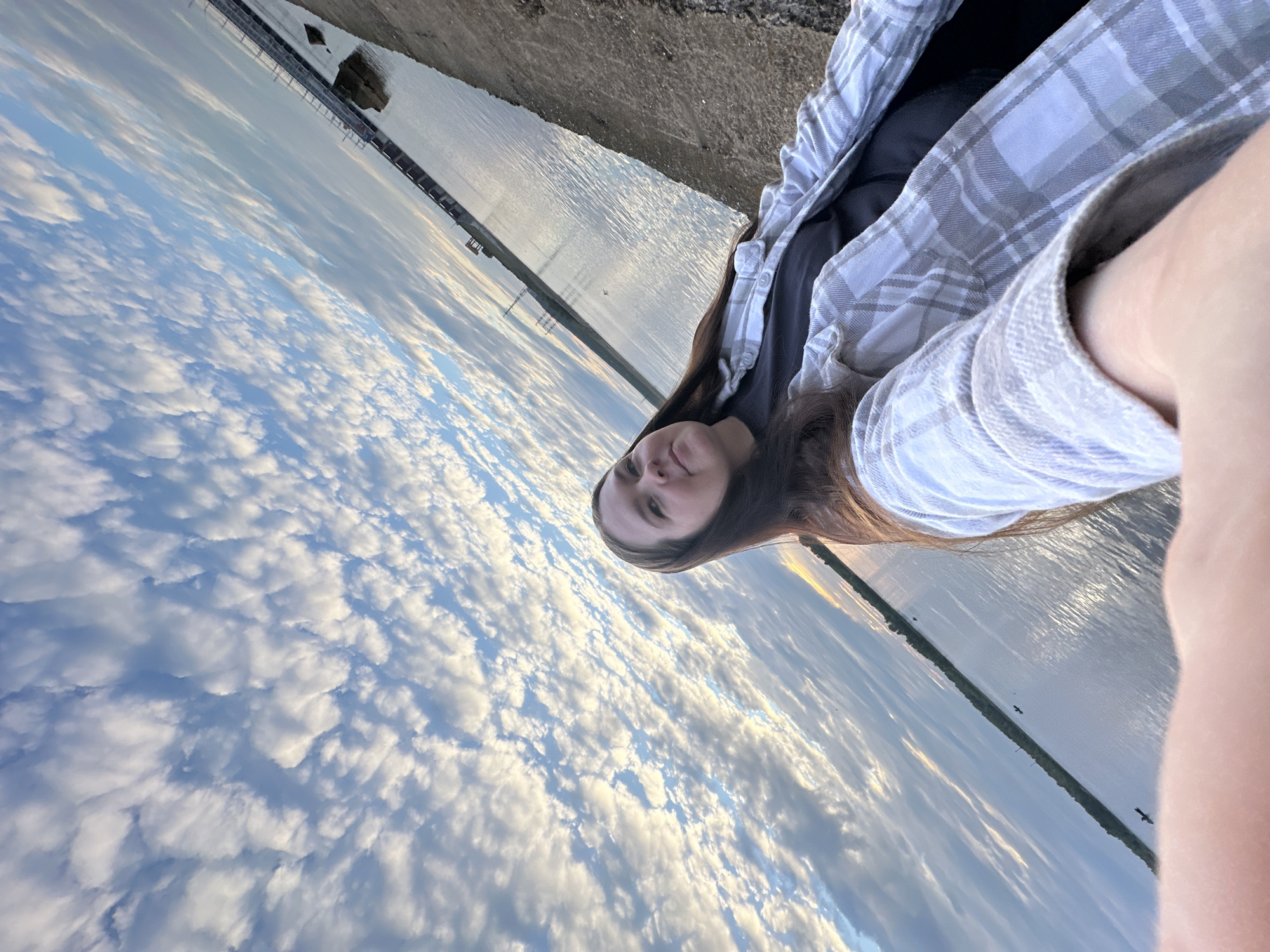A high-angle, wide selfie shows a woman with long, dark hair and fair skin. She is wearing a gray t-shirt and an unbuttoned, light-colored plaid shirt. She is looking slightly up and to her right, with a neutral expression. The background is dominated by a dramatic sky filled with wispy, white clouds against a light blue sky. A body of water, possibly a river or lake, stretches horizontally across the image, with a narrow strip of land on the far side featuring some vegetation and structures, including power lines and a dam or industrial facility. A concrete or gravel embankment is visible in the foreground, on which the woman is standing or sitting. The scene appears to be outdoors, possibly at dusk or dawn, given the lighting conditions in the sky.