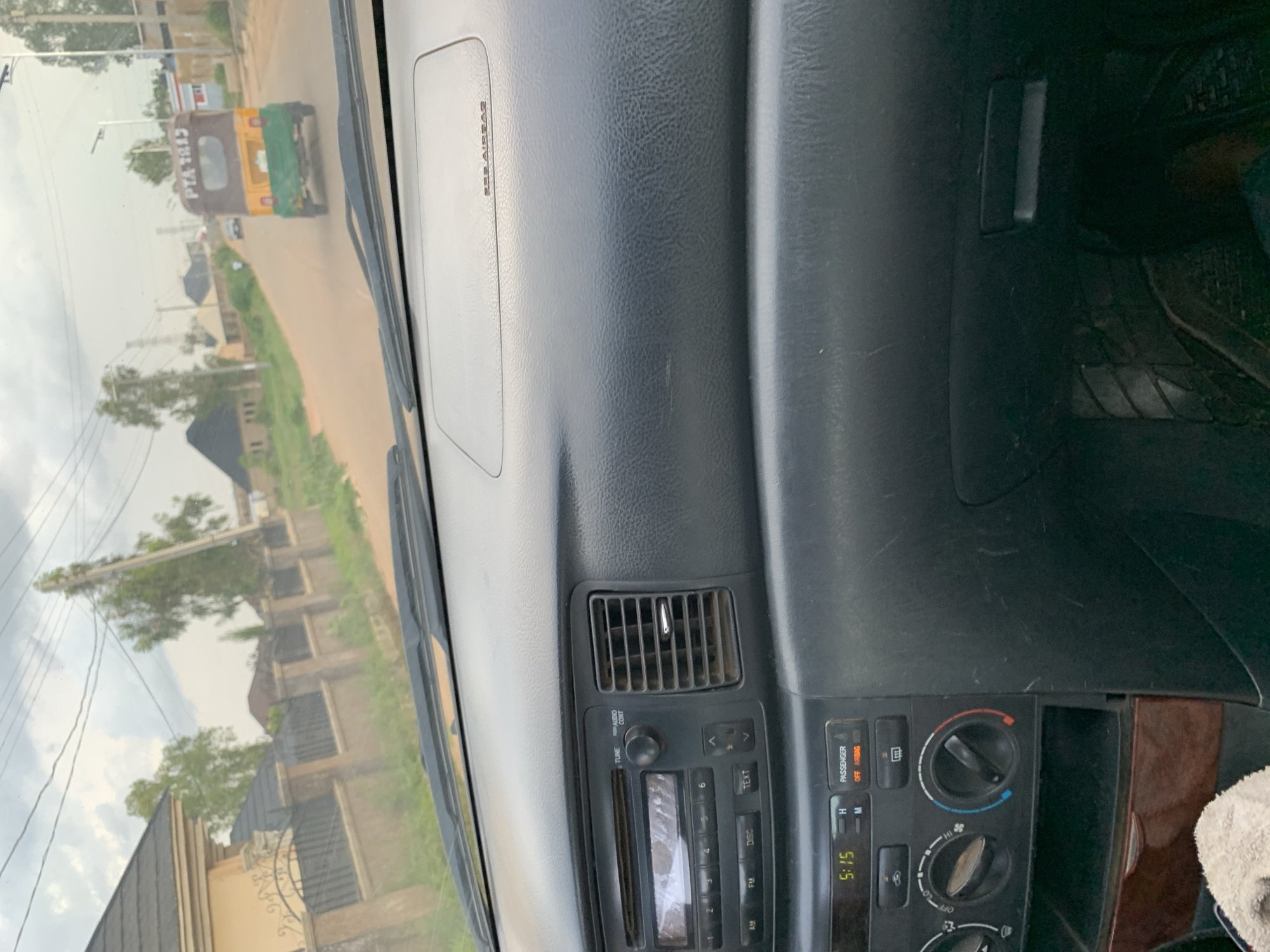 The image, captured from inside a vehicle, shows a street scene in Jos, Nigeria. The dashboard of the car is visible, including the radio, air conditioning controls, and an "SRS AIRBAG" indicator. Outside the car, the road stretches ahead, with buildings and vegetation lining the sides. A yellow and green auto-rickshaw, often referred to as a "Keke Napep" or "Keke," is visible in the distance, with the license plate "PTA 1113" clearly legible. The sky is overcast, suggesting cloudy weather. The overall environment appears to be a suburban or semi-urban area.