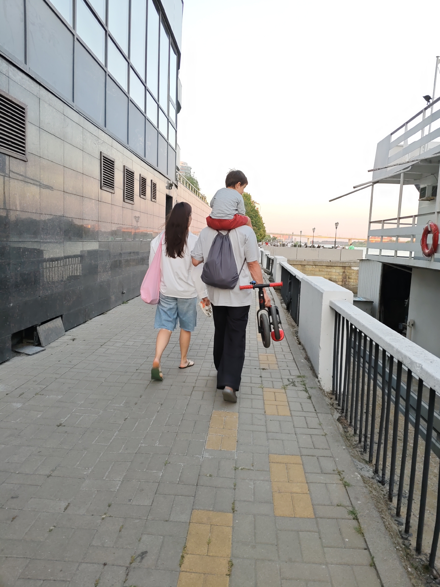 Two adults and a child are walking on a paved sidewalk next to a body of water and a building with reflective windows. The person in front is a woman wearing denim shorts and a white long-sleeved shirt, carrying a pink tote bag. She is walking to the left of the frame. To her right and slightly behind her is a man wearing black pants and a white t-shirt. He is carrying a young child on his shoulders, and the child is wearing red shorts and a gray t-shirt. The man is also carrying a small bicycle. A white boat with a red life preserver is docked to the right of the walkway, separated by a black metal railing. The building to the left has a dark facade with horizontal vents. The sky is overcast and pale. The scene appears to be a waterfront promenade in Rostov-on-Don, Russia.