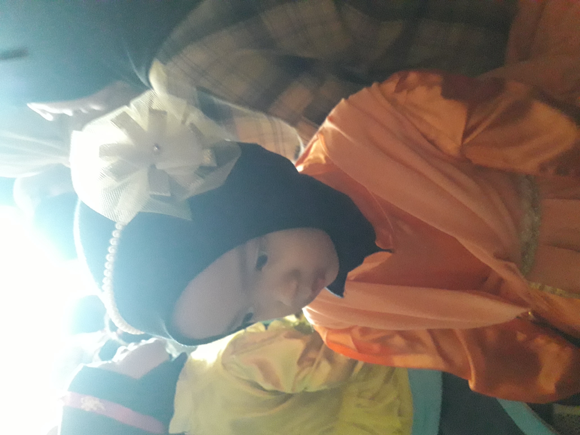 The image depicts a close-up of a young child wearing an orange garment and a black hijab adorned with a white bow and pearls. The child is looking down and to the left, with their mouth slightly open. In the background, other individuals are partially visible, also wearing similar attire, suggesting a cultural or religious gathering. The lighting is bright and appears to be from an artificial source, creating a strong glare in the upper right portion of the image. The overall scene is informal and likely captured in an indoor setting. The image is from Cankuang, Indonesia.