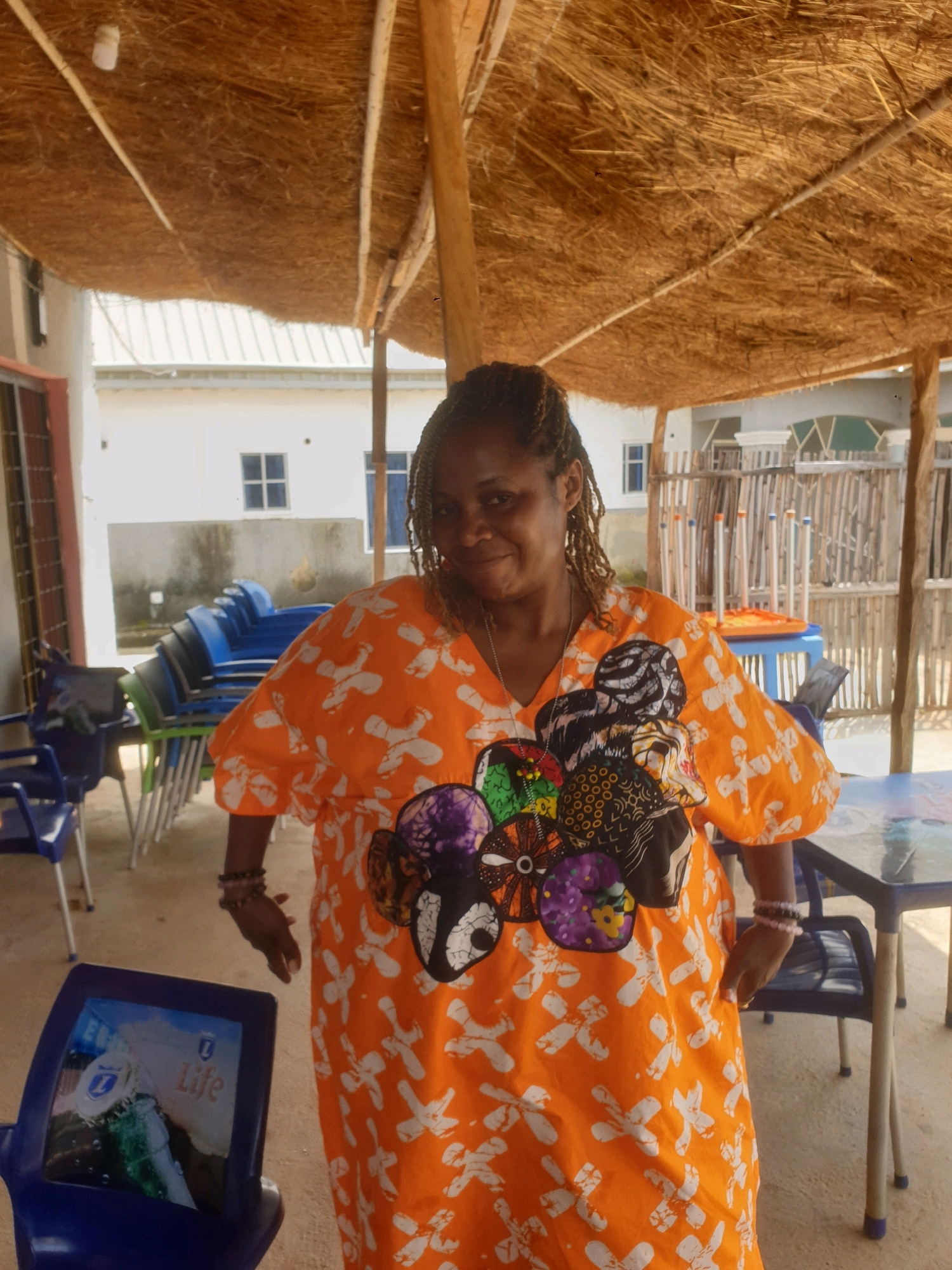 In Mangu Town, Nigeria, a woman stands in an outdoor seating area. She wears a vibrant orange dress with a white pattern and a colorful, circular design on the chest. Her hair is styled in braids, and she is adorned with bracelets. Behind her, several blue chairs are stacked, and a table with a glass top is visible. The seating area is covered by a thatched roof supported by wooden poles, and a fence made of bamboo-like sticks is in the background. The buildings in the distance appear to be white with windows. The overall scene suggests a casual, outdoor dining or gathering space.