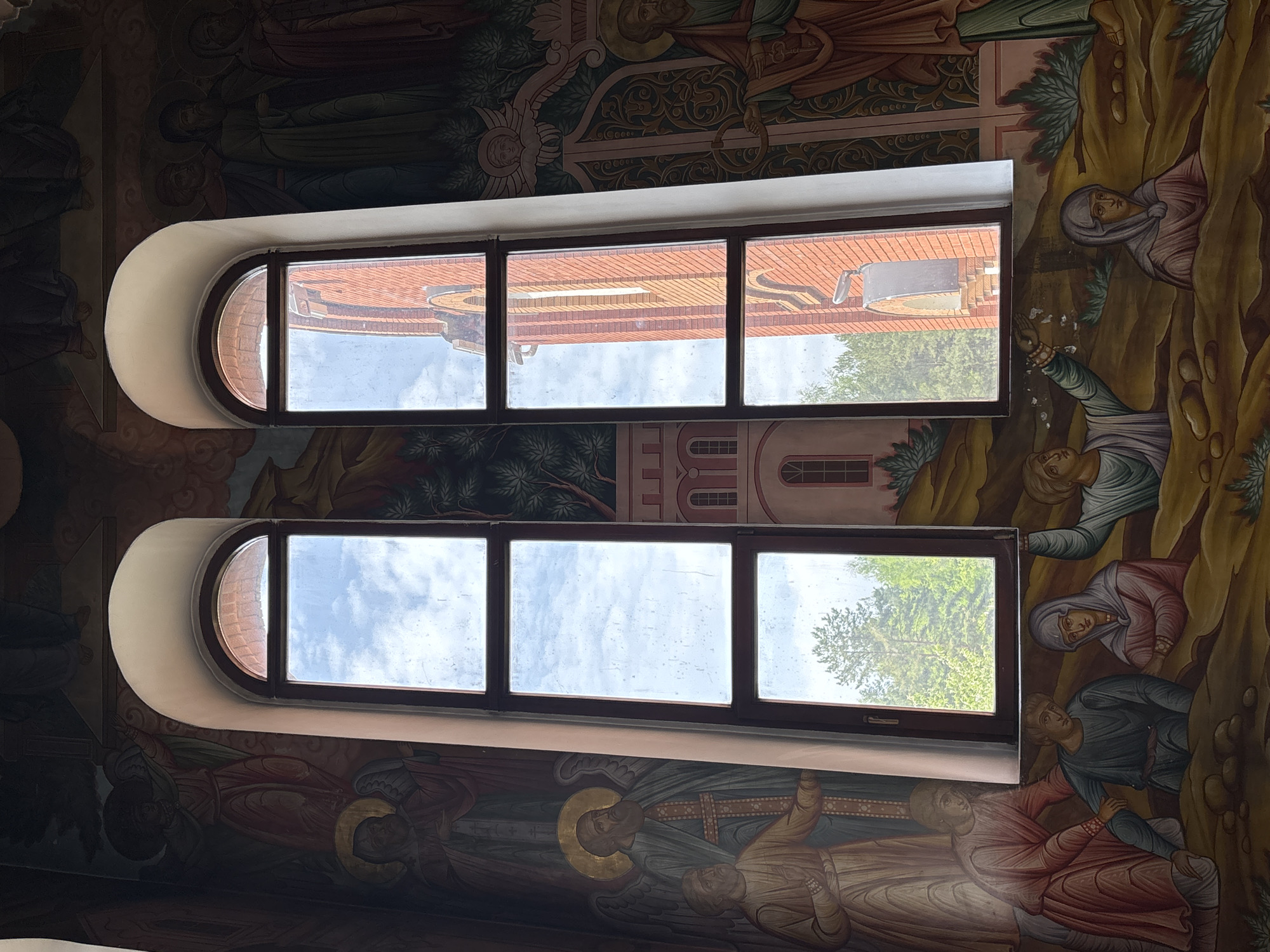The photograph shows two arched windows in a building interior. The walls around the windows are decorated with religious frescoes. Through the upper window, part of a building with a red tile roof is visible, while the lower window shows trees and sky. The location is Chaikovsky, Russia.