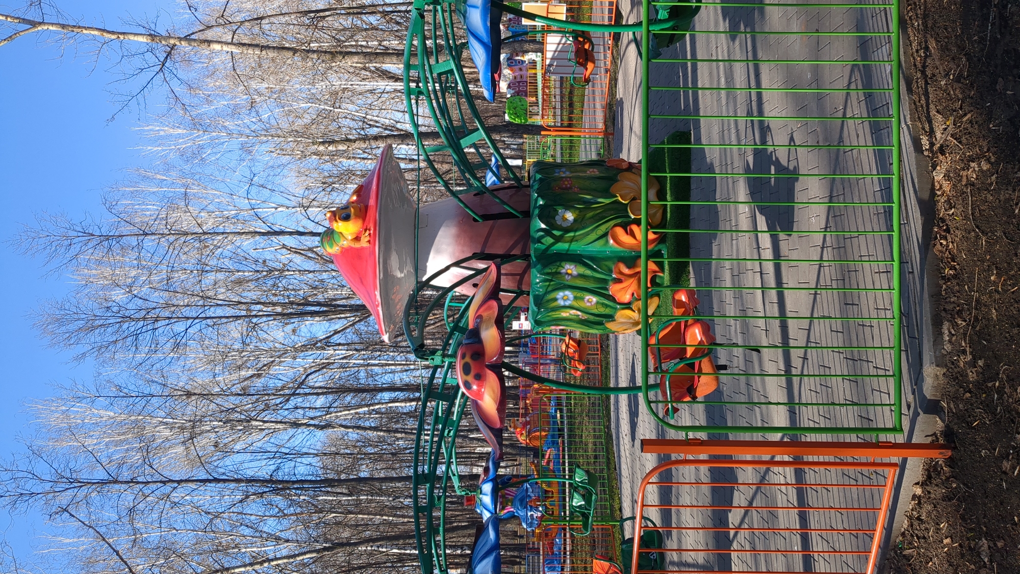 The image displays a colorful children's amusement park ride in an outdoor setting, likely in Ulyanovsk, Russia. The central feature of the ride is a large, mushroom-shaped structure with a vibrant red cap, on top of which sits a green and yellow frog character. Below this, several individual ride cars are visible, including one designed as a red and black ladybug. The ride's base is adorned with green panels decorated with white, yellow, and orange floral and leaf patterns. The individual seats for riders appear to be orange.
The ride area is enclosed by a green metal fence. Adjacent to this, there is a paved pathway made of light grey interlocking bricks. Shadows from the fence and other structures are cast distinctly on the pavement, indicating bright, direct sunlight. Beyond the paved area, a strip of dark soil covered with scattered leaves and debris is visible, bordered by a curb. Another orange metal fence is present in the foreground, suggesting an entrance or boundary of the park.
The background features numerous deciduous trees with bare branches, set against a clear, bright blue sky. This absence of foliage suggests the image was taken during late autumn, winter, or early spring. The weather appears sunny and clear. No people are visible in the scene, and there is no discernible text. The overall atmosphere is quiet, highlighting the colorful, whimsical nature of the children's attraction.