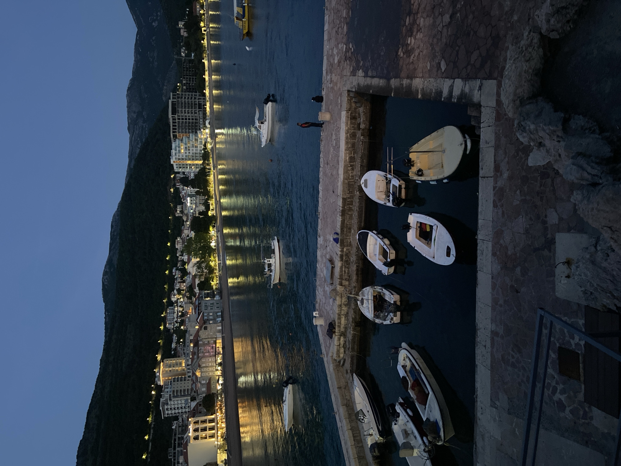The image shows a nighttime view of the harbor area in Petrovac centar, Montenegro. Along the left side of the frame, a hillside is visible with lights from buildings along the coast reflecting in the water. Docked boats are seen in a small marina to the right. A person wearing red can be seen walking along the edge of a dock. The sky above is a dark blue.