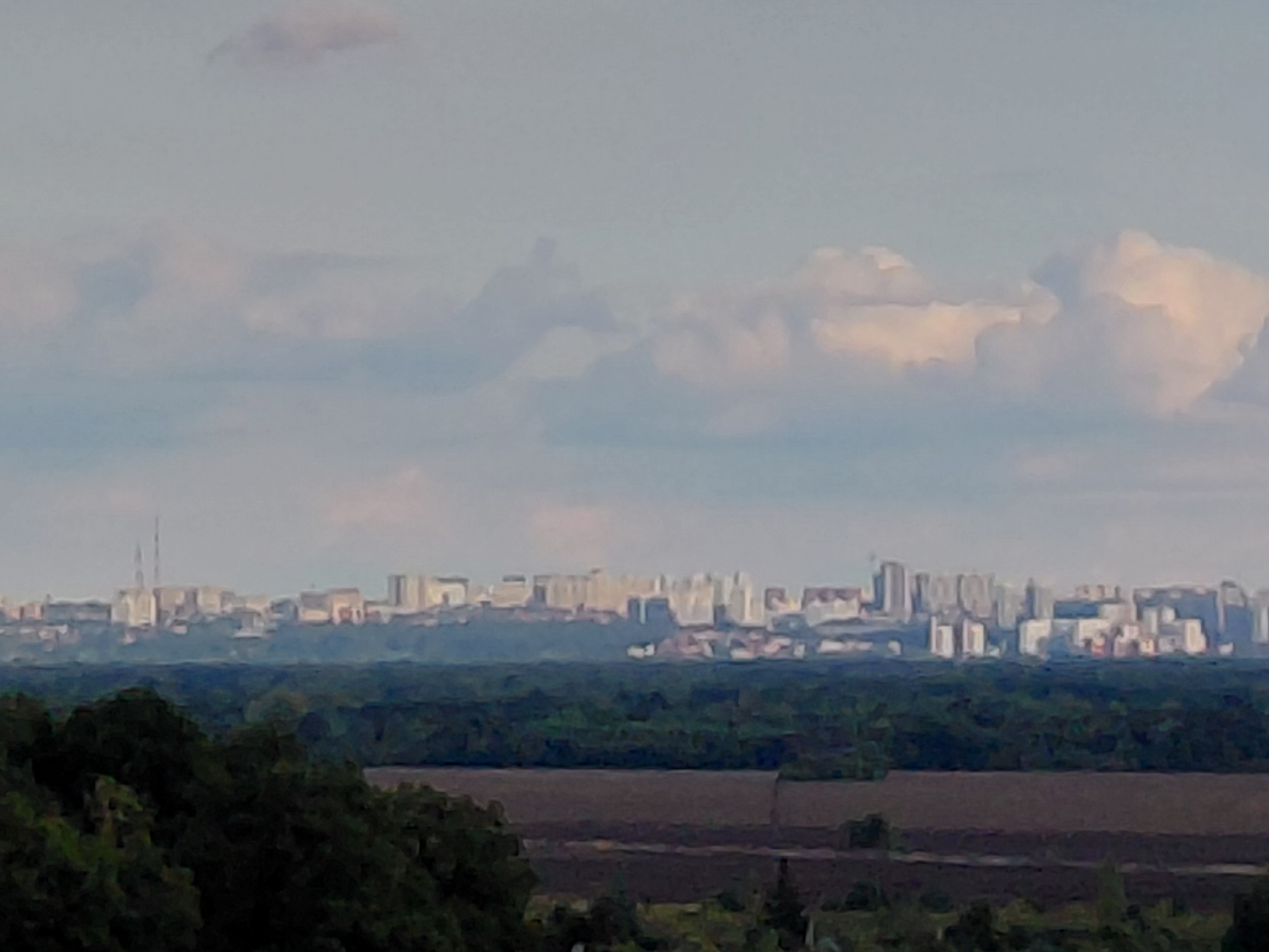 This is a distant, eye-level, landscape photograph of a city skyline in Samara, Russia, under a hazy, overcast sky. The foreground features the dark green foliage of trees and shrubs, with a field of tilled brown earth visible beyond. In the mid-ground, a dense line of dark green trees forms a natural barrier. The background showcases a sprawling urban landscape with numerous light-colored, multi-story buildings, including apartment blocks and what appear to be industrial structures. Two tall, slender towers, possibly communication towers or chimneys, are prominent on the left side of the city. The overall impression is one of a vast, populated area seen from a distance, with a transition from natural to urban environments.