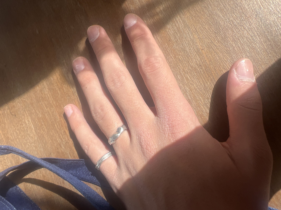 The image displays a human left hand, palm down, resting on a light-colored wooden surface. The hand exhibits light-toned skin with visible texture. Two silver-colored rings adorn the fourth digit: a thin, twisted band is positioned distally on the proximal phalanx, while a thicker, irregularly contoured band is located proximally to it on the same phalanx. The fingernails are unpolished, showing natural appearance, visible cuticles, and minor surface irregularities. Direct sunlight illuminates the scene, casting distinct shadows from the hand onto the wooden surface. A dark blue fabric strap is partially visible in the upper left quadrant of the image. The location is Hof Ashkelon Regional Council, Israel.