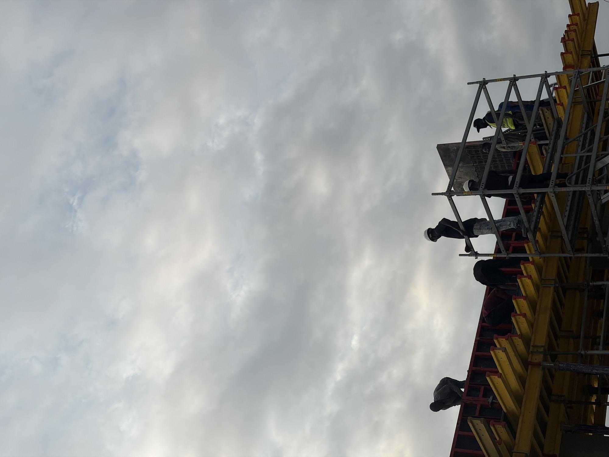 In Alaguntan, Nigeria, several construction workers are actively engaged on a multi-level scaffolding structure under a cloudy sky. The scene is dominated by yellow and red formwork for concrete casting. On the upper level, a worker in dark clothing is bent over, seemingly inspecting or adjusting the formwork. To their left, another worker in dark clothing is also bent over the formwork. Further to the right, a worker wearing a white hard hat and dark clothing stands on the scaffolding. On a lower section of the scaffolding, to the right, a group of workers are visible. One worker is wearing a neon yellow safety vest and a dark cap, appearing to be directing or observing. Another worker next to them is wearing a white hard hat. The sky is overcast with diffused light, suggesting either early morning or late afternoon. The overall impression is a busy construction site actively working on a concrete structure.