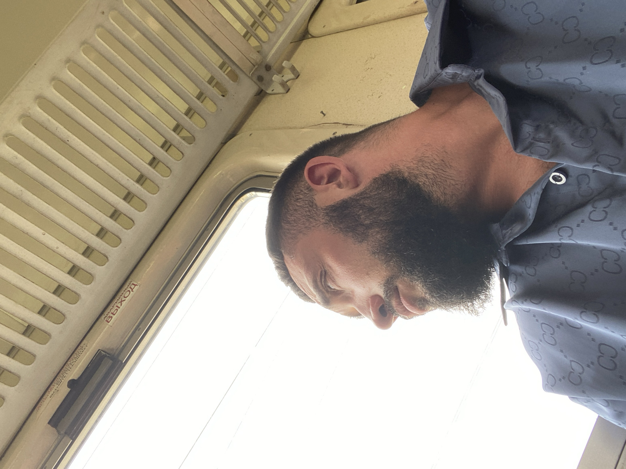 A man with a beard and dark hair is captured in a low-angle, close-up shot, seemingly taken from within a mode of transport like a train or bus, given the presence of a slatted overhead panel and a window. The man is looking down, his face partially obscured by his beard, with his eyes closed or looking downwards, suggesting a moment of rest or introspection. He is wearing a dark shirt with a repeating pattern that appears to be a logo, possibly a designer brand.
Visible text in the image includes Russian words. On a strip near the window, "АВАРИЙНЫЙ ВЫХОД" (Emergency Exit) is clearly legible, along with the word "ВЫХОД" (Exit) again on a red sticker. This indicates the location is likely a public transportation vehicle operating in a Russian-speaking region. The lighting suggests it might be daytime, with bright light coming from the window. The overall atmosphere is candid and personal.