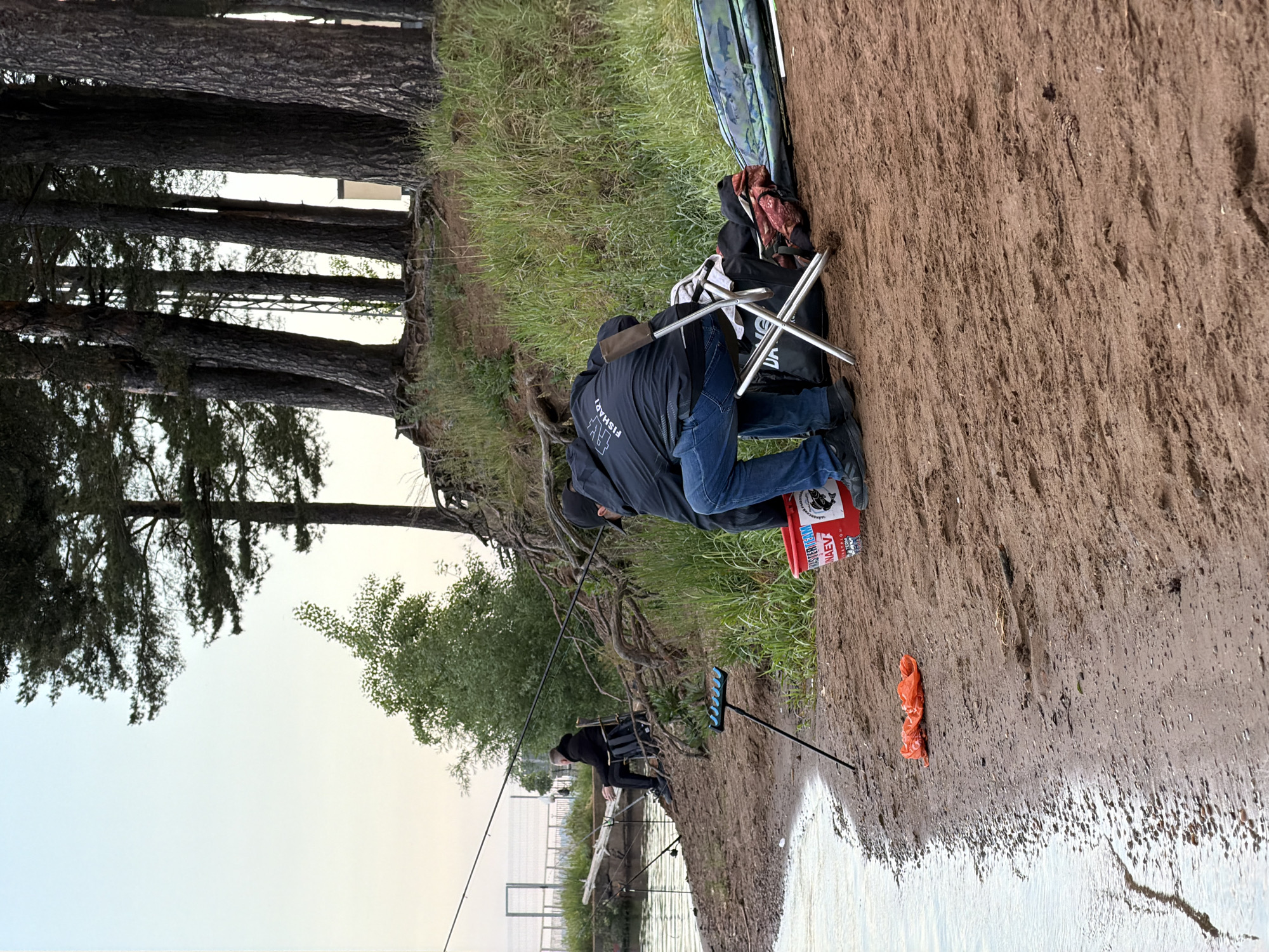 The image captures an outdoor scene of two individuals fishing on the sandy bank of a body of water, likely a river or lake. The scene is set in Chaikovsky, Russia.
In the foreground, a person is seen from behind, seated on a portable metal camping chair. They are wearing a dark jacket with "FISHART" visible on the back, and blue jeans. Several fishing rods are set up in rod holders along the water's edge, indicating active fishing. A red bucket with the text "MASTERWORM" and partially visible "MAEBA" is placed near their feet. Adjacent to them, a black bag and a green camouflage bag, possibly containing fishing gear, rest on the sand. An orange plastic item lies on the ground closer to the water.
Further down the bank to the left, another person is also engaged in fishing, seated on a fishing box or stool, with their own rod extended into the water.
The environment features a steep, sandy and muddy bank with exposed tree roots. Tall, slender pine trees dominate the left side of the frame, providing a natural backdrop. The water is calm, reflecting the soft, overcast sky, which suggests either early morning or late afternoon. In the distance across the water, a faint structure resembling a fence or distant pier can be discerned. The overall impression is one of peaceful recreation and focus on the activity of fishing.