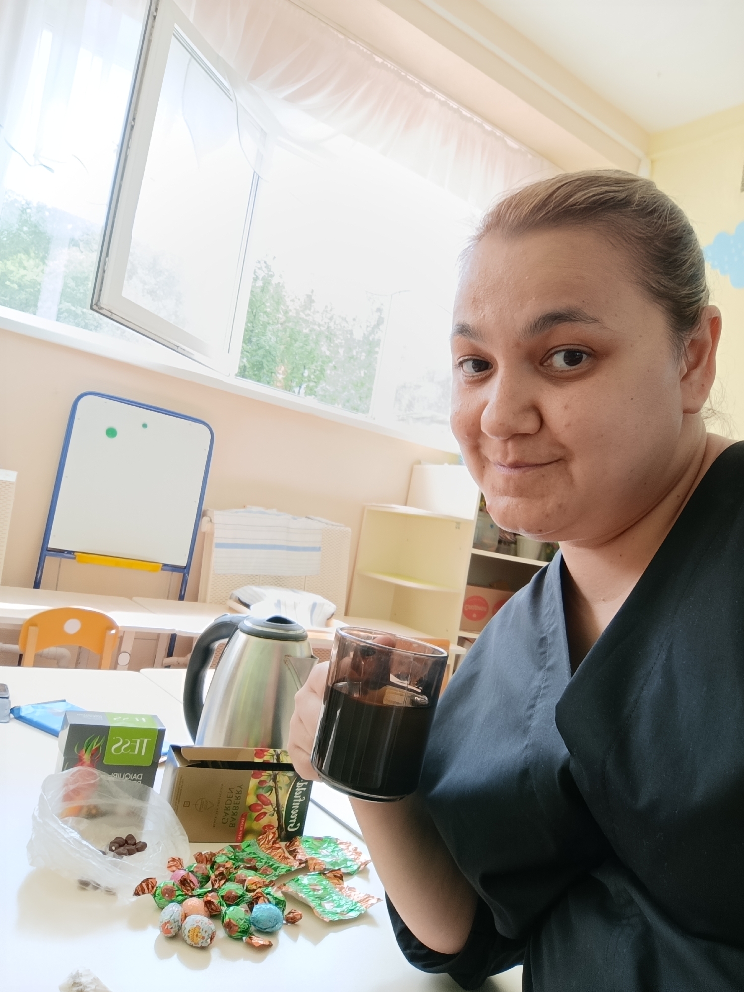 A woman stands inside a brightly lit room in Chaikovsky, Russia, holding a glass of dark liquid, possibly tea or coffee. A silver electric kettle, a box of Tess tea, a box labeled "Sibderevo Bukreevskie Sweets," a bag of chocolate candies, and unwrapped assorted wrapped candies are on the white table in front of her. A whiteboard on a blue frame and an open window with sheer curtains are behind her. The woman is wearing a dark-colored uniform or top.