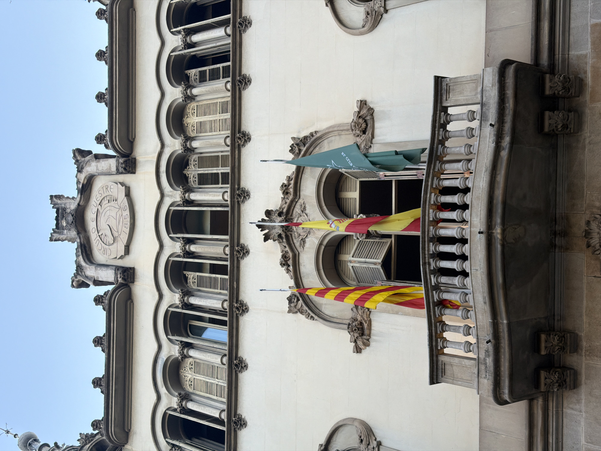 This is a low-angle, full shot of a building's facade in Barcelona, Spain, specifically the Círculo Ecuestre building. The building's architecture is ornate, with multiple arched windows that feature decorative columns and shutters. A prominent balcony with a stone balustrade juts out from the second story. Three flags are displayed from this balcony: the Catalan flag on the left, the Spanish flag in the center, and a green flag with a white emblem on the right. Above the balcony, the facade is adorned with decorative stonework and a circular emblem with the inscription "CIRCULO ECUESTRE 1856." The sky above is clear and pale blue, suggesting it is daytime. There are no people visible in the image.