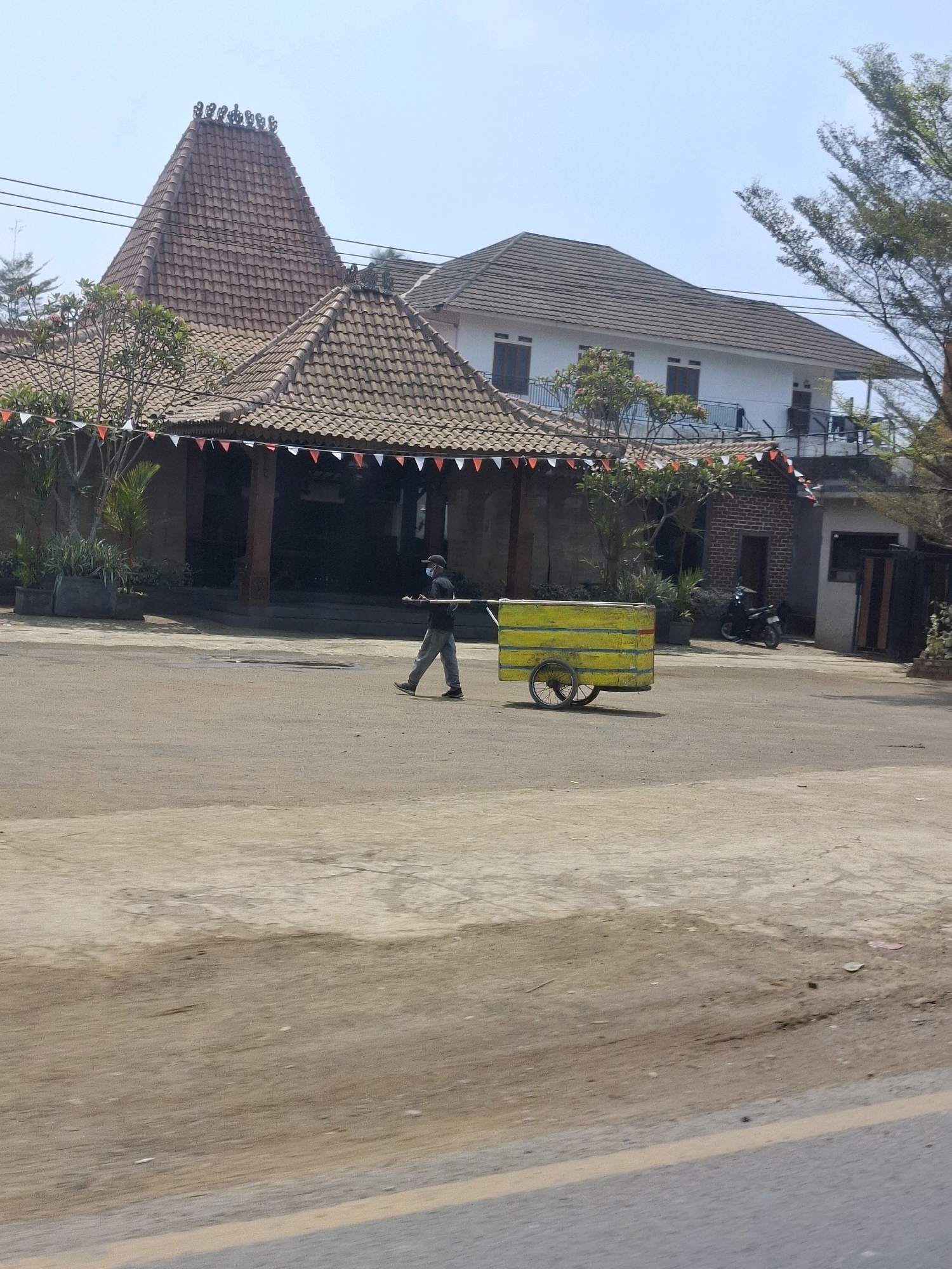 A person wearing a face mask and casual clothing pushes a yellow cart with two wheels. The cart appears to be made of wood with horizontal yellow stripes. The person is walking on an asphalt-like surface in front of a building complex with traditional Indonesian architecture, featuring prominent tiled roofs and decorative elements. Red and white triangular flags are strung along the roofline of one of the buildings, suggesting a possible celebration or national holiday. In the background, there is a white two-story building and a brick structure. A motorcycle is parked near the brick building. The environment suggests an outdoor setting in a semi-urban or rural area. The image is taken during daylight with a clear sky.