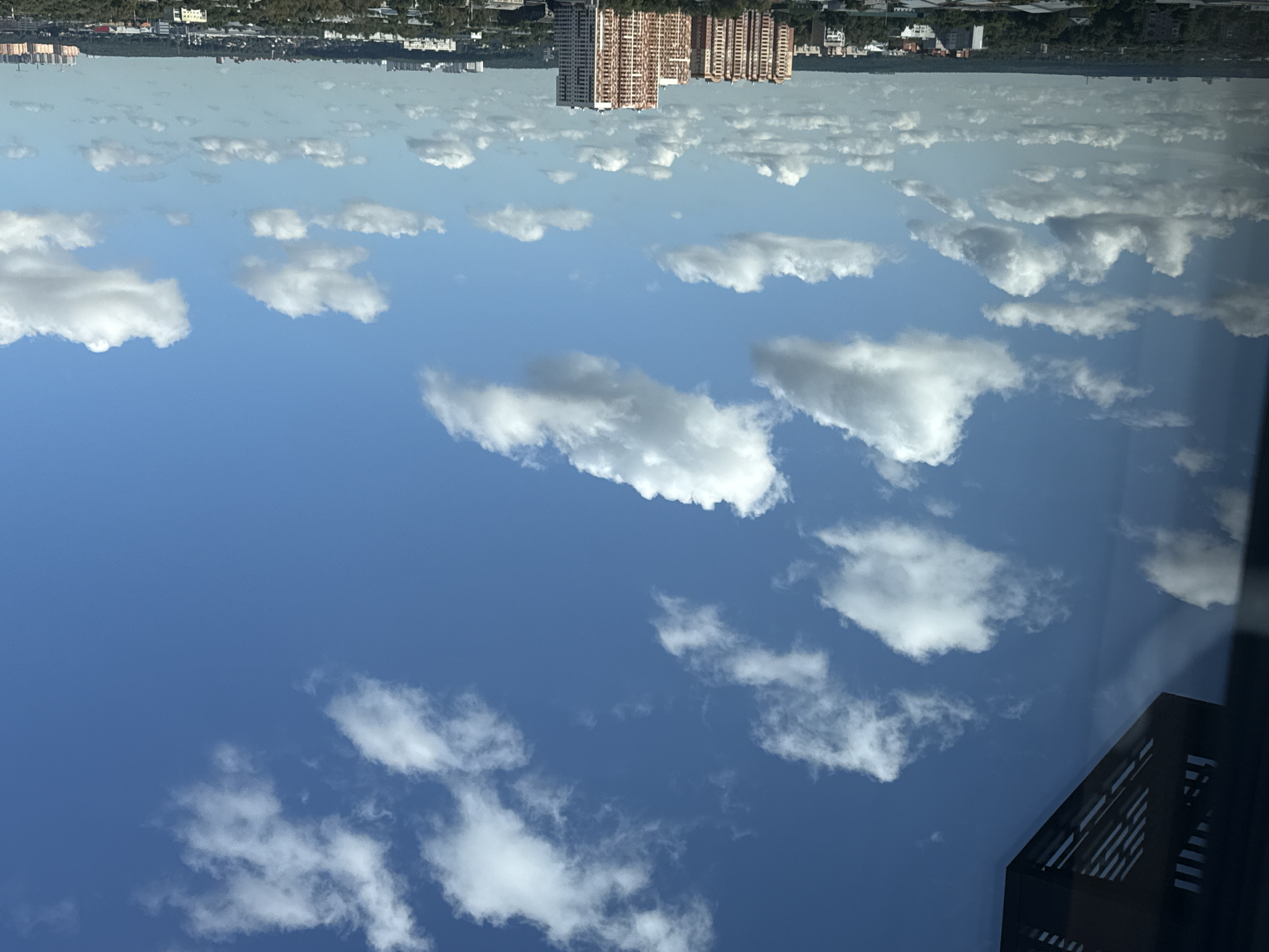 The image shows a sky with scattered white clouds over a cityscape. In the foreground, a dark, angular structure with horizontal slats is visible on the left side, possibly a part of a building or a balcony. The middle ground displays a dense collection of trees and various buildings, including multi-story residential blocks with balconies and patterned facades, and some smaller structures. In the distance, a faint horizon is visible with more buildings and a green expanse of trees, suggesting a sprawling urban area. The sky is a vibrant blue, with fluffy white clouds of varying sizes and densities dispersed across it. The lighting suggests it is daytime, likely mid-morning or afternoon, given the brightness and clear visibility of the cityscape. The overall scene appears to be an aerial view looking out from an elevated position within the city of Lyubertsy, Russia.
