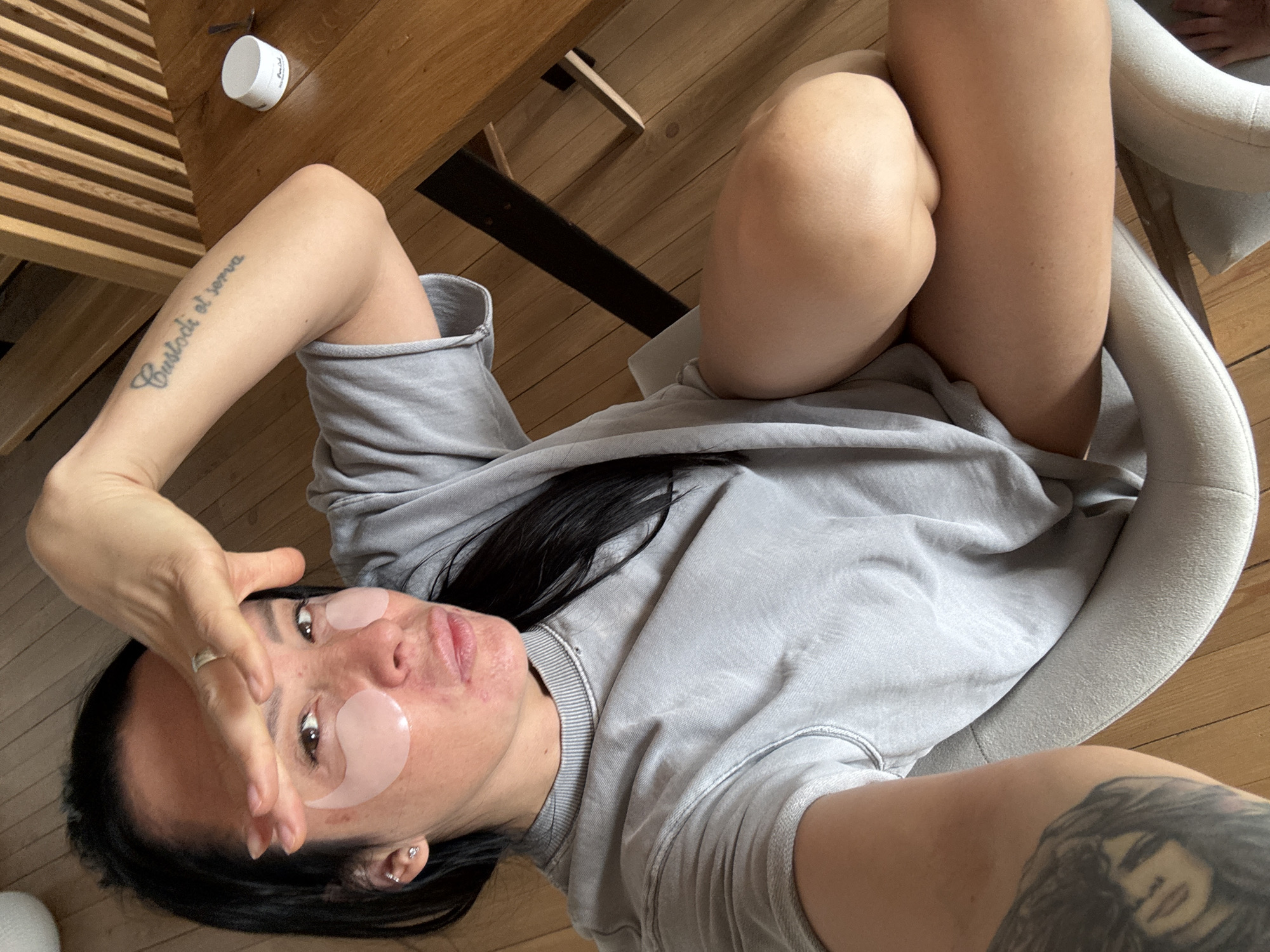 An overhead shot depicts a person, likely a woman, with dark hair, lying down and looking directly at the camera. She is wearing a light grey, long-sleeved shirt and grey shorts or sweatpants, and has two pink eye patches placed under her eyes, suggesting a skincare routine. Her left arm is bent, with fingers partially obscuring her forehead, and a silver ring is visible on her left hand. A tattoo on her left forearm clearly reads "Custodi et serva". Another tattoo, depicting a portrait of a face, is visible on her right arm.
The setting appears to be indoors, characterized by light-colored wooden flooring and wooden furniture, possibly a table or chair, with visible slats. A small white jar or container with partially legible text, possibly "Purity," rests on the wooden surface near her left arm. Parts of a light-colored, upholstered chair or seat are visible on the right side of the frame. The lighting suggests daytime. The person's expression is neutral and relaxed. The image does not contain specific visual cues to confirm the location as Chaikovsky, Russia.