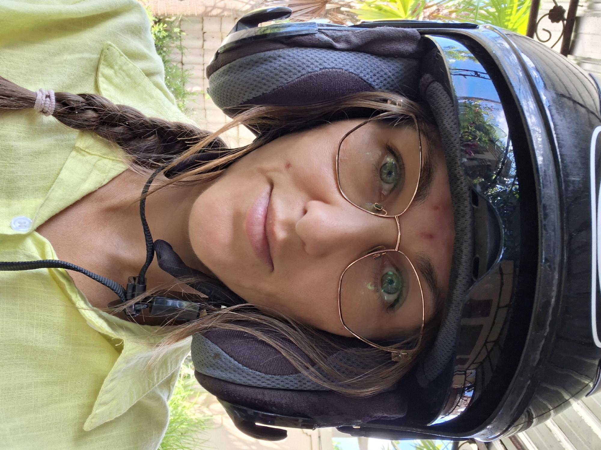 A woman is shown wearing a black motorcycle helmet with tinted visor. She has light skin, green eyes, and is wearing gold-rimmed glasses. Her hair is brown and partially braided. A small microphone attached to a cord is visible near her collar. She is wearing a light green shirt. The helmet's visor reflects an outdoor scene with vegetation. The photo was taken in the Ubud District of Indonesia.