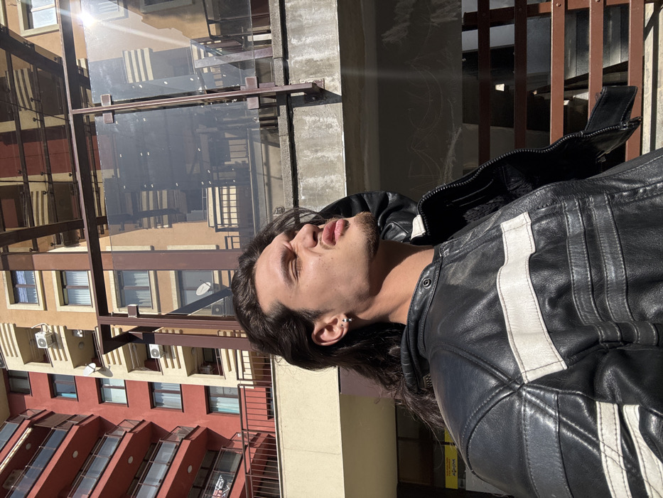 An outdoor portrait in Sofia, Bulgaria, features a male individual from the chest up, positioned against an urban backdrop. He has long, dark hair, a short beard, and wears a silver hoop earring in his left ear. He is dressed in a black leather jacket with prominent white horizontal stripes and a visible hood. His face is turned to the right, eyes squinted, and lips pursed, indicating a reaction to direct sunlight. His left hand is partially visible near his temple. The immediate background includes a concrete ledge and a structure composed of brown metal framing and clear glass panels, which reflects elements of the surrounding environment. Beyond this, two multi-story buildings are visible: one predominantly red with numerous balconies on the left, and another light brown structure on the right. Both buildings display multiple windows and external air conditioning units. The scene is brightly illuminated by natural sunlight, casting strong light and shadows.
