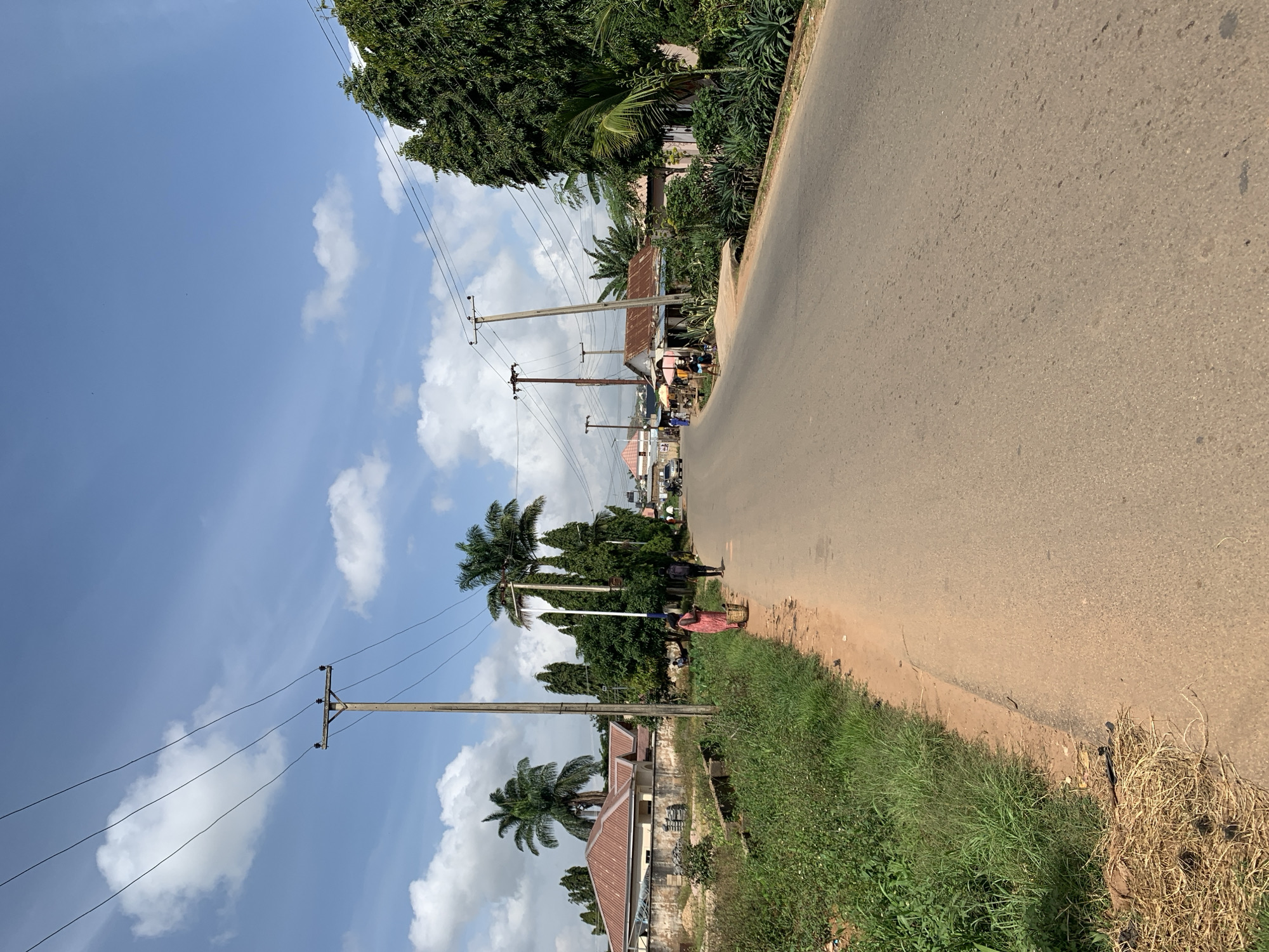 A rural road stretches into the distance under a partly cloudy sky. Several utility poles with wires are visible along the roadside, supporting the power grid. The road is lined with lush green vegetation, including tall palm trees and dense shrubs. On the left side of the road, houses with reddish-brown roofs are nestled behind the foliage. On the right, a few simple structures with corrugated metal roofs are present, suggesting a residential or commercial area. Several individuals are seen walking along the road. One person in the foreground on the left is carrying a basket. Further down the road, other pedestrians and a vehicle are visible, indicating ongoing activity. The overall scene depicts a typical day in a Nigerian town.