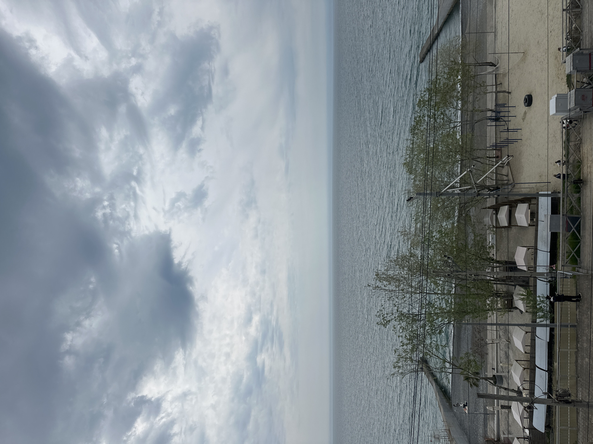 This image captures a wide view of a quiet coastal area in Sochi, Russia, under an overcast sky. The photograph is oriented vertically, but when rotated horizontally, it reveals a vast expanse of greyish-blue water on the right, likely the Black Sea, meeting a distant horizon line.
The sky on the left side of the image is dominated by thick, grey, and white clouds, indicating an overcast or partly cloudy day with diffused light.
Below the horizon, a narrow strip of sandy and pebbly beach runs along the coastline. On the beach and an adjacent elevated area, numerous white sun loungers or cabanas are visible, arranged in neat rows. Scattered across the scene are several deciduous trees with sparse, light green foliage, suggesting early spring. A dark tire rests on the sand, and some metal structures, possibly for exercise or play, are also present.
A long, dark breakwater or pier extends into the water from the left side of the beach. The foreground features a boardwalk or paved promenade running parallel to the water, lined with utility poles, electrical wires, and various white and grey utility boxes. One person, dressed in dark clothing, stands by a railing on the promenade in the lower right corner, looking towards the sea.
The scene appears calm and sparsely populated, with no significant activity taking place beyond the lone individual. The overall mood is subdued due to the cloudy weather. No legible text is visible in the image.