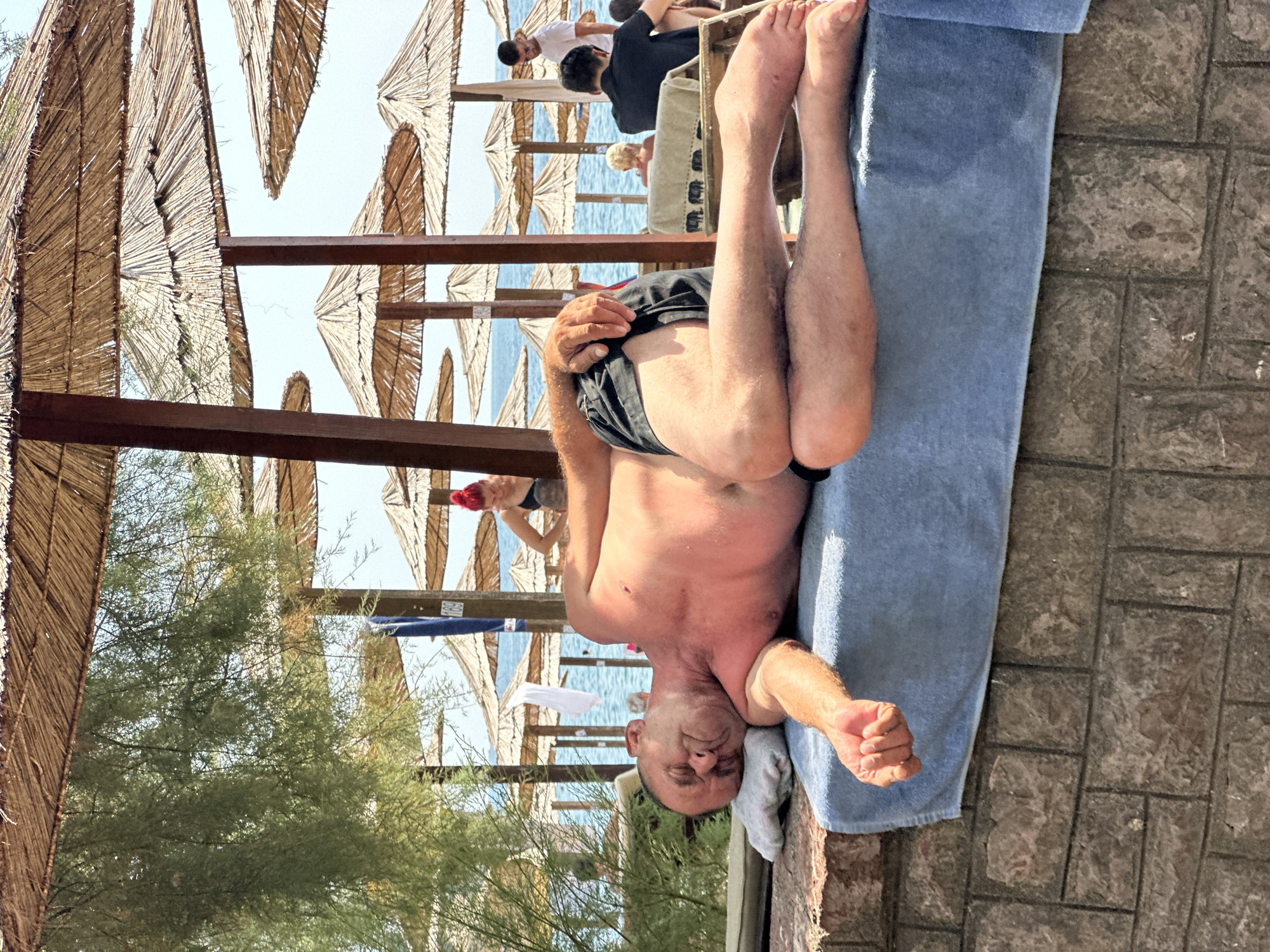 A middle-aged man with tanned skin is lying on his back on a blue towel, with his head resting on a light gray pillow. He is wearing black swim trunks and appears to be asleep or resting with his eyes closed. His left arm is extended to his side, while his right arm is bent at the elbow, with his hand resting on his chest. The man is lying on a raised platform with a brick base, covered by the towel.
In the background, a beach scene is visible with numerous thatched umbrellas providing shade. Beyond the umbrellas, the blue sea stretches out under a clear sky. Several other people are present on the beach, including a woman with bright red hair visible under an umbrella, and men standing and sitting in the background. The environment suggests a sunny day at a beach resort, likely in Budva, Montenegro, given the context.
