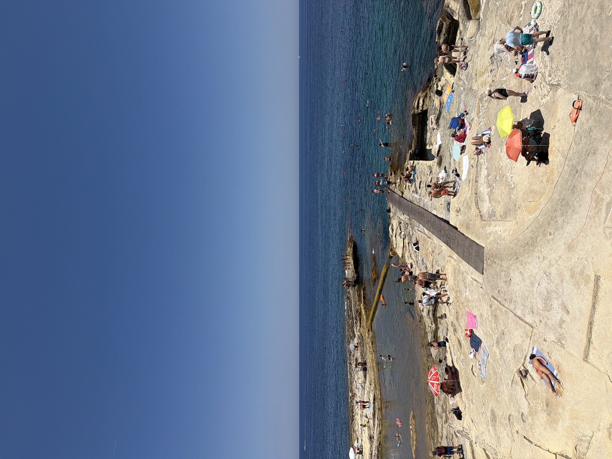 This is a high-angle, wide shot of a rocky shoreline in Sliema, Malta, on a clear, sunny day. The deep blue Mediterranean Sea occupies the upper two-thirds of the frame, with a faint line of the horizon separating it from the clear blue sky. A small white sailboat is visible far out at sea.
The lower third of the image depicts a popular sunbathing and swimming area carved into the natural rock formations. Several groups of people are scattered across the sun-baked rocks. In the foreground, a person lies on a towel, sunbathing. A little further back, two colorful umbrellas, one orange and one yellow, provide shade for a small group of people. Various towels and bags are spread out on the rocks.
A wooden walkway extends from the rocky shore into the water, leading to a small island-like rock formation where more people are gathered. In the shallow, clear water near the shore, several individuals are swimming or wading. Some pink inflatable floats are visible in the water. The rocky coastline is characterized by its light-colored, textured surface, with some scattered vegetation. The overall scene suggests a lively and popular spot for recreation and relaxation.