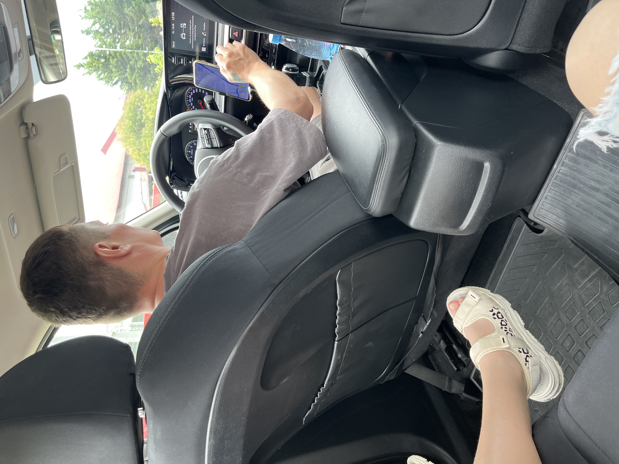 A man is driving a car in Sochi, Russia. He is wearing a grey t-shirt and has short brown hair. He is looking at his phone, which is mounted on the dashboard. A woman's leg, wearing a white sandal, is visible in the foreground. The car's interior is dark and appears to be a modern vehicle with a touch-screen display and leather seats. Outside the car, trees and buildings are visible, suggesting an urban or suburban setting. The weather appears to be overcast.