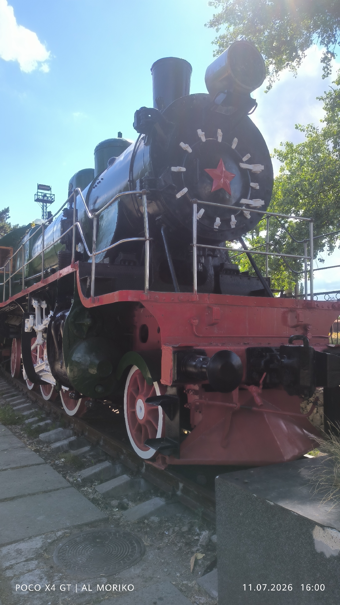 The image shows a close-up, low-angle view of a vintage black and red steam locomotive, likely a Soviet-era model, on display. The locomotive is positioned on railroad tracks, with concrete sleepers and gravel visible. A bright red star emblem is centered on the front of the locomotive, surrounded by white protrusions. The locomotive features a large smokestack, a prominent headlight, and large red wheels with white rims. Green foliage from trees is visible in the upper right corner, and the sky is blue with some clouds. A metal railing runs along the side of the locomotive. The bottom of the image contains text overlaying the scene: "POCO X4 GT | AL MORIKO" on the left and "11.07.2026 16:00" on the right. There are no people visible in the image. The setting appears to be an outdoor public space, possibly a park or a museum exhibit, located in the city of Bender, Moldova.