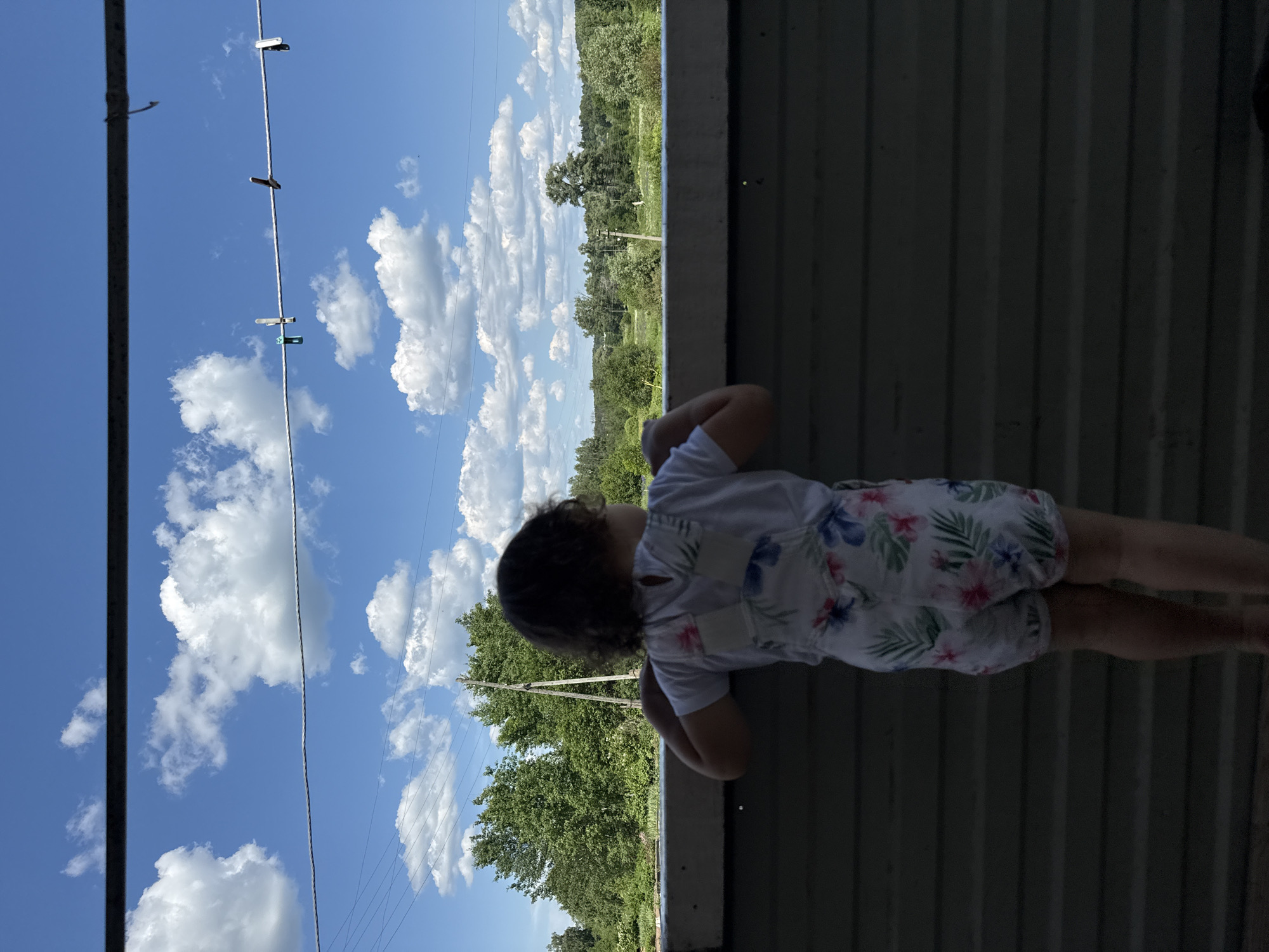 A young child with curly brown hair, wearing a white t-shirt and floral print overalls, stands facing away from the camera, looking out at a landscape. The child's hands rest on a dark-colored railing. The background reveals a bright blue sky filled with fluffy white clouds. A clothesline with several clothespins is strung horizontally across the upper portion of the image. Below the sky, a line of green trees and vegetation extends across the horizon, with a few power poles visible amongst them. The scene appears to be set outdoors, possibly on a porch or balcony, overlooking a rural or semi-rural area in the city of Уральское, Russia.