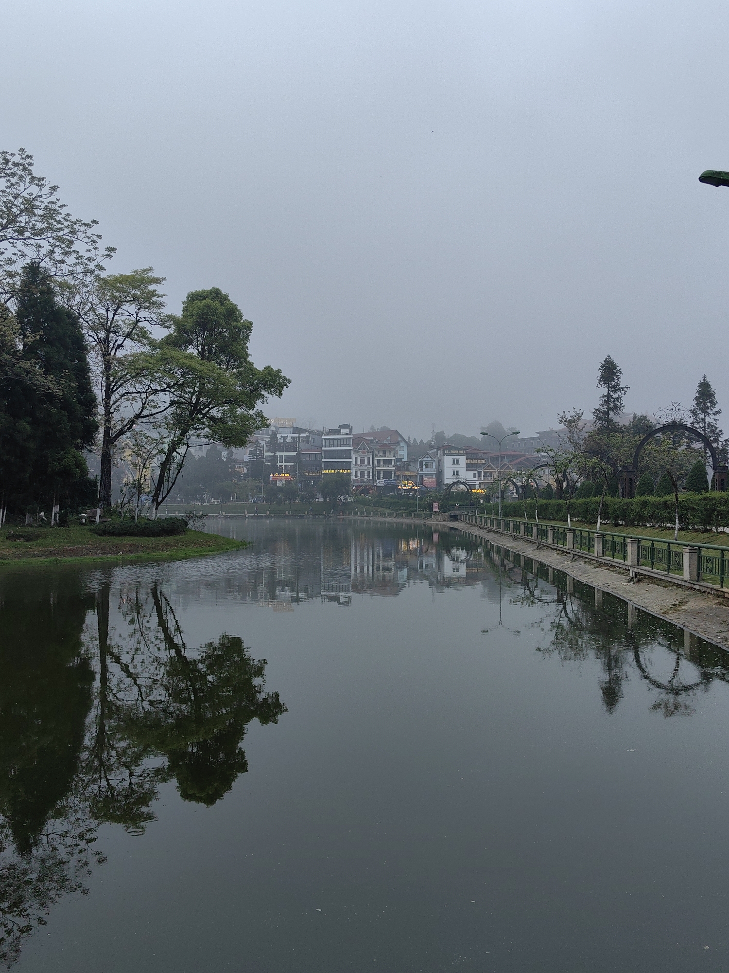 The image depicts a panoramic view of Sa Pa Lake in Sa Pa, Vietnam, under heavy mist and overcast conditions. The sky is uniformly light grey, limiting visibility of distant features. The placid, dark-surfaced lake occupies the majority of the lower frame, exhibiting slight ripples and reflecting the surrounding environment.
On the left shoreline, a small landmass extends into the lake, featuring several deciduous and coniferous trees, some with green foliage and others with sparse branches. Their detailed reflections are visible in the water.
The right shoreline is bordered by a paved embankment and a green metal railing with stone posts. Behind this, a landscaped area includes manicured hedges, smaller trees, and two ornate metal archways. A green-hooded streetlamp is partially visible on the far right.
Across the lake, a dense cluster of multi-story buildings, primarily commercial or hotel structures, forms the background. These buildings display varied architectural styles, numerous windows, and illuminated signage, with text such as "NEN SA PA HOTEL" and "LE THUY HA" discernible on certain facades. The upper sections of these structures and the mountainous terrain beyond them are significantly obscured by the pervasive mist. No individuals or specific actions are visible within the scene.