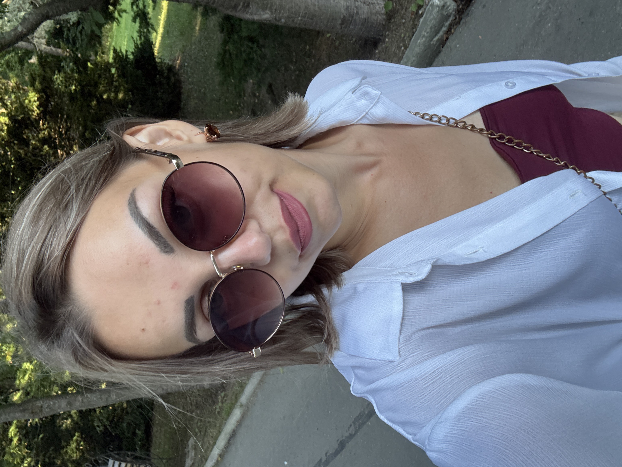 A young woman with shoulder-length blonde hair and light skin is captured in a selfie outdoors. She is wearing large, round, rose-tinted sunglasses, a white open-collared shirt, and a dark red top underneath. She has light makeup on, including pink lipstick. A small, amber-colored earring is visible on her right ear. The background suggests a park or wooded area with trees and a paved path. The scene is lit by natural daylight, casting soft shadows. The overall impression is casual and relaxed. It is not possible to determine the specific location within Yuzhno-Sakhalinsk, Russia, from the visual information provided.