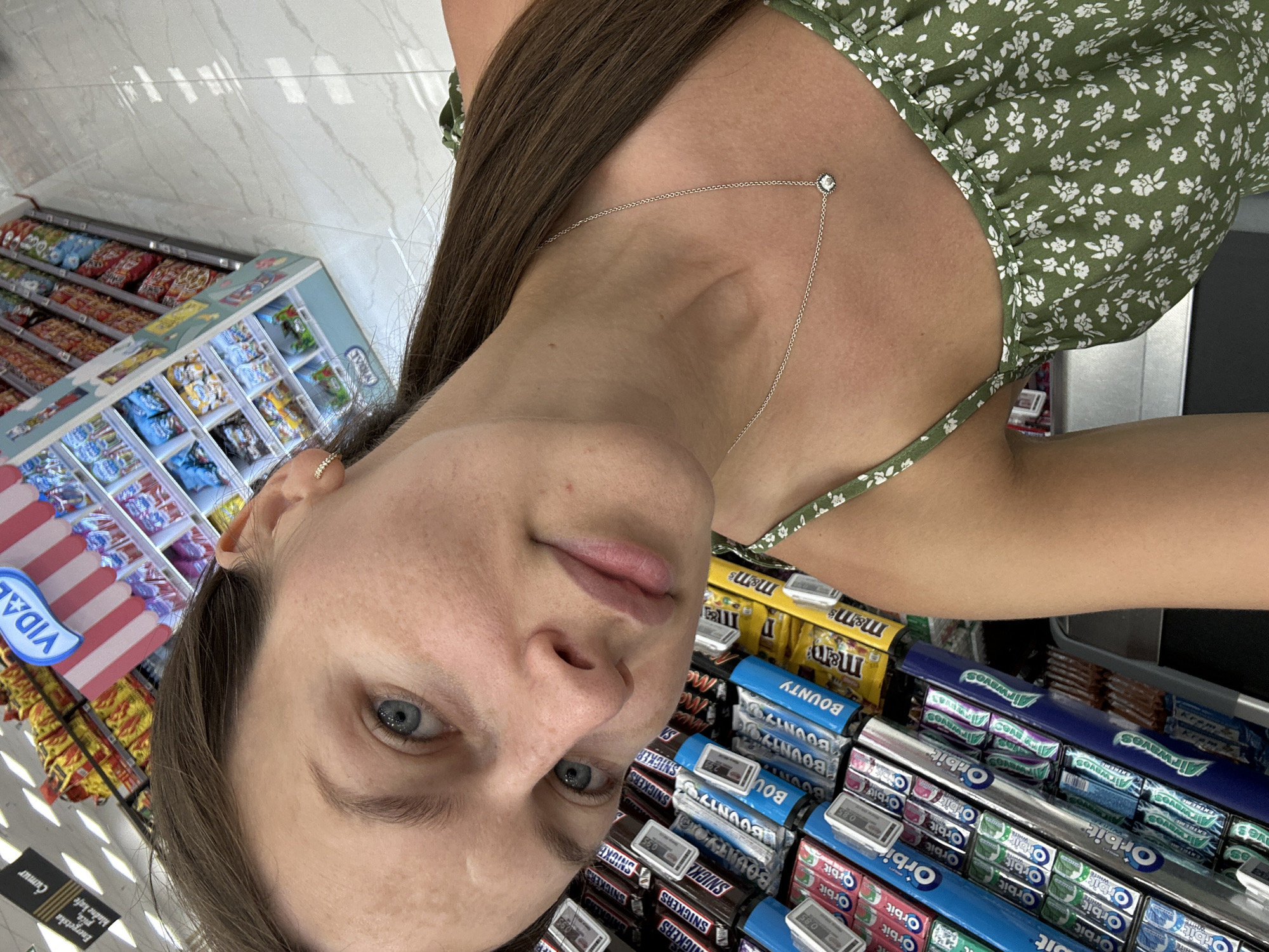 A woman is taking a selfie in a supermarket aisle. She is wearing a green floral dress and a silver necklace. The aisle is filled with candy bars, gum, and other snacks. The supermarket is located in Donji Pobori, Montenegro.